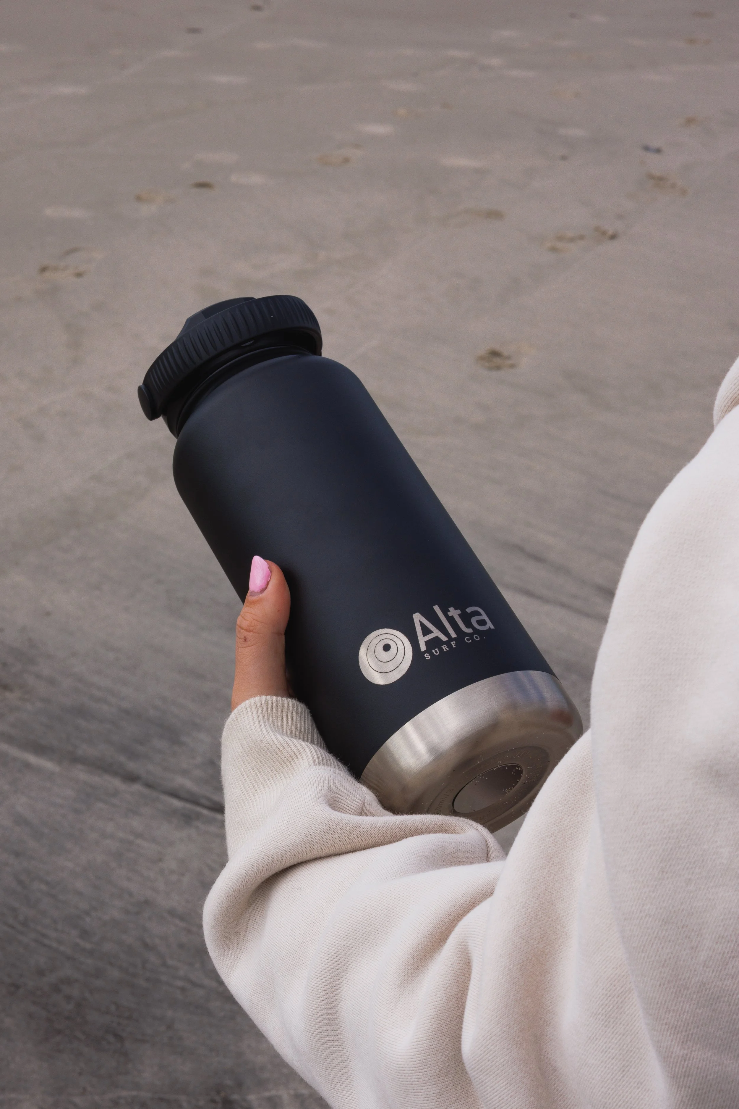 A person wearing a white sweater holding a black insulated tumbler with the brand name Alta Surf Co. on a paved surface, with some footprints visible in the background.