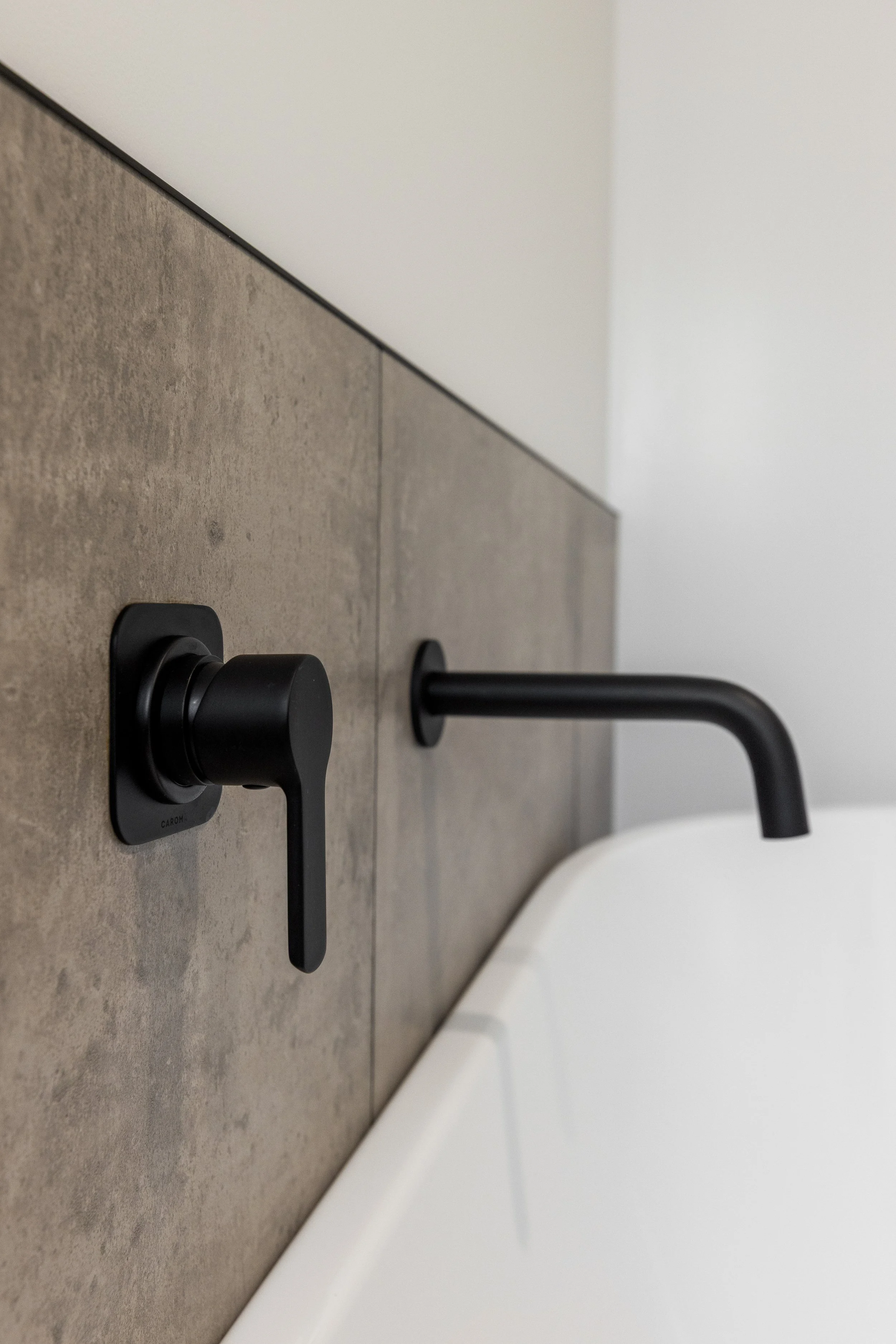 Close-up of a black soap or sanitizer dispenser on a gray tiled wall above a white sink in a modern bathroom or washroom.