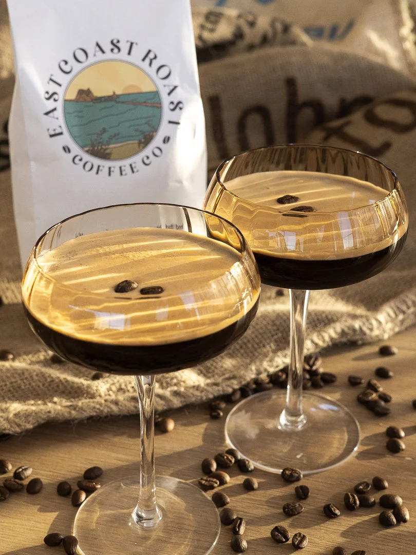 Two coupe glasses filled with espresso martinis garnished with coffee beans, with a bag of coffee beans and a coffee bag in the background.