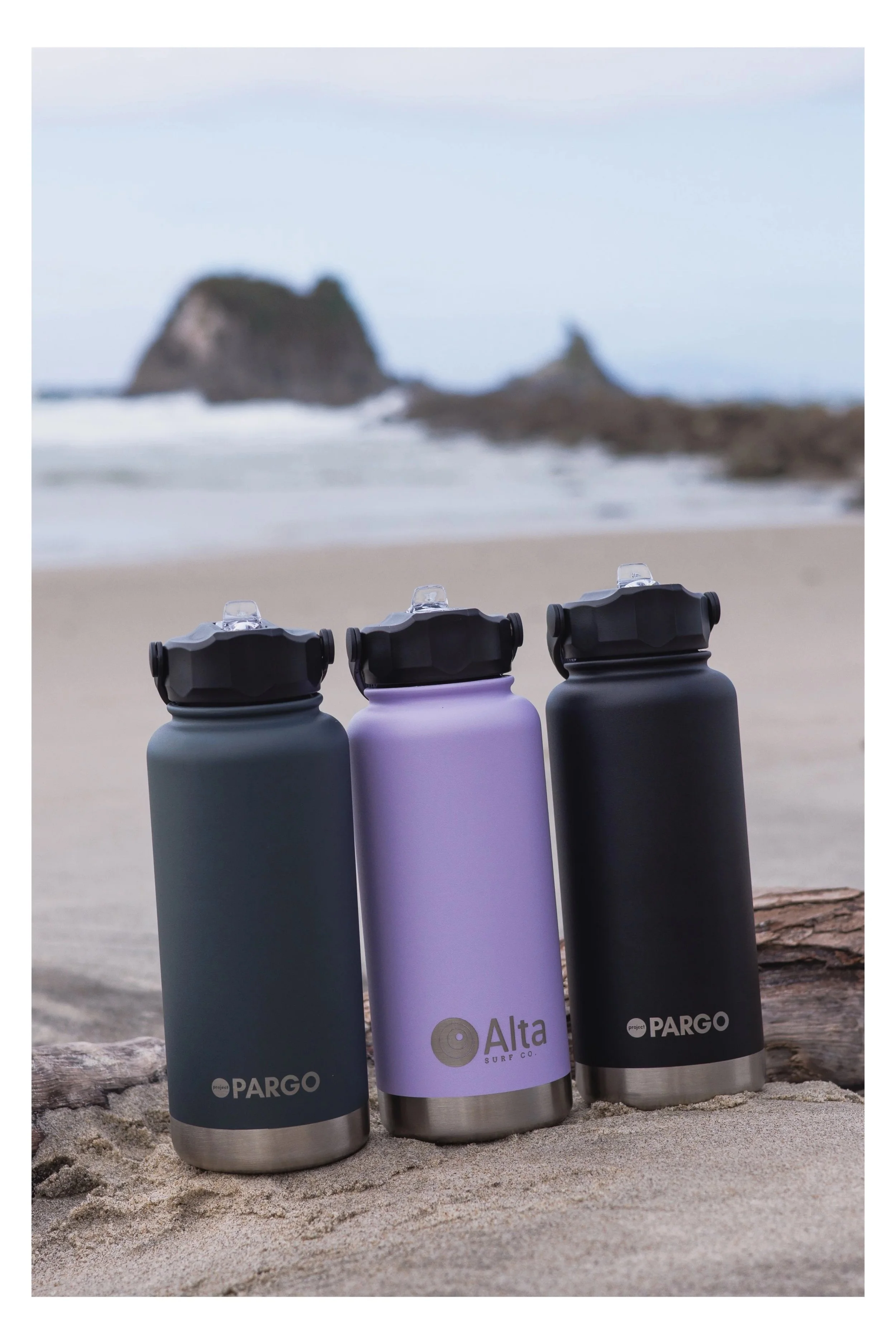 Four reusable water bottles with black caps and stainless steel bottoms, labeled 'Pargo' and 'Alta Surf Co.', rest on a sandy beach with a blurred rocky island or cliff in the background.