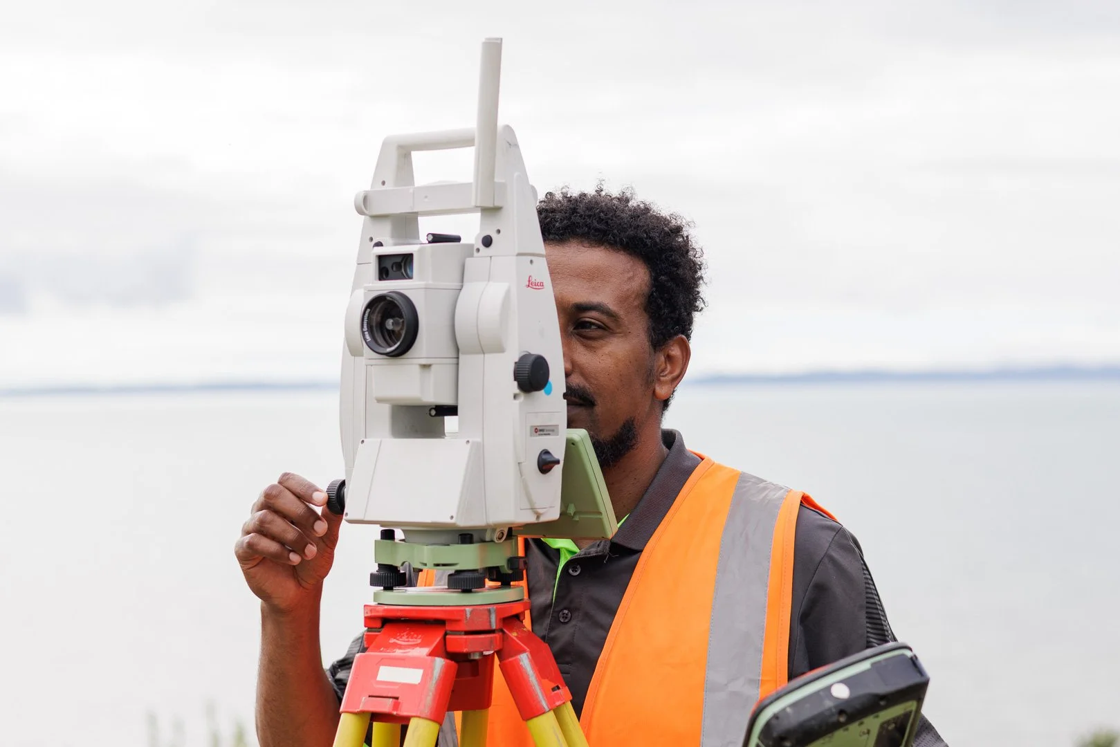 Man operating a theodolite or surveying instrument outdoors on a cloudy day, wearing an orange safety vest and holding a device in his hand.
