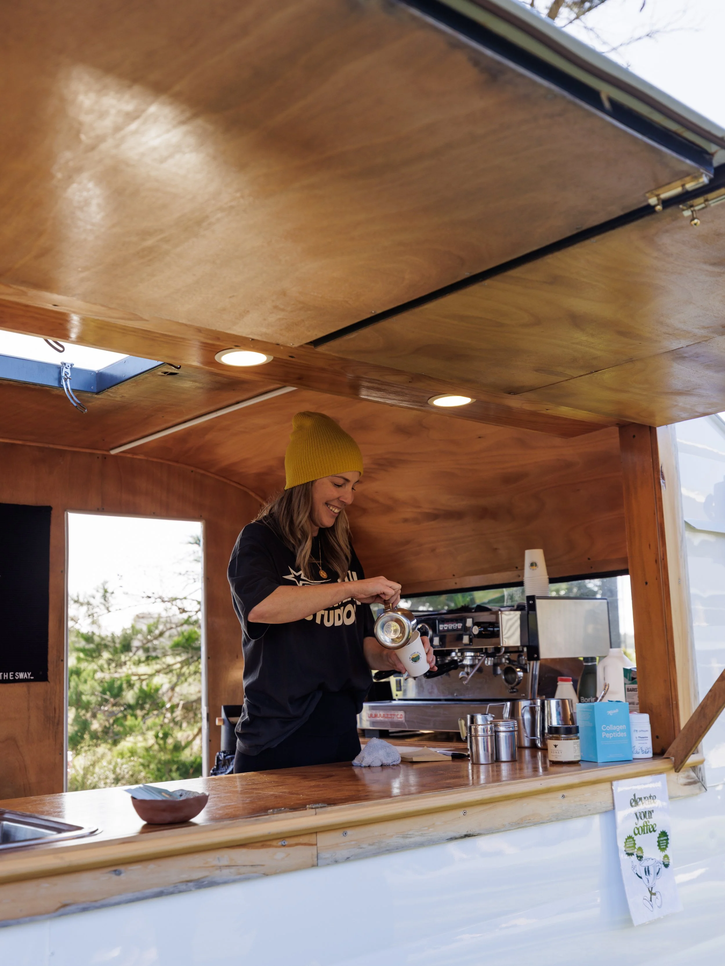 A woman with a yellow beanie and black shirt preparing coffee at a wooden coffee stand outdoors, with a smiling expression.
