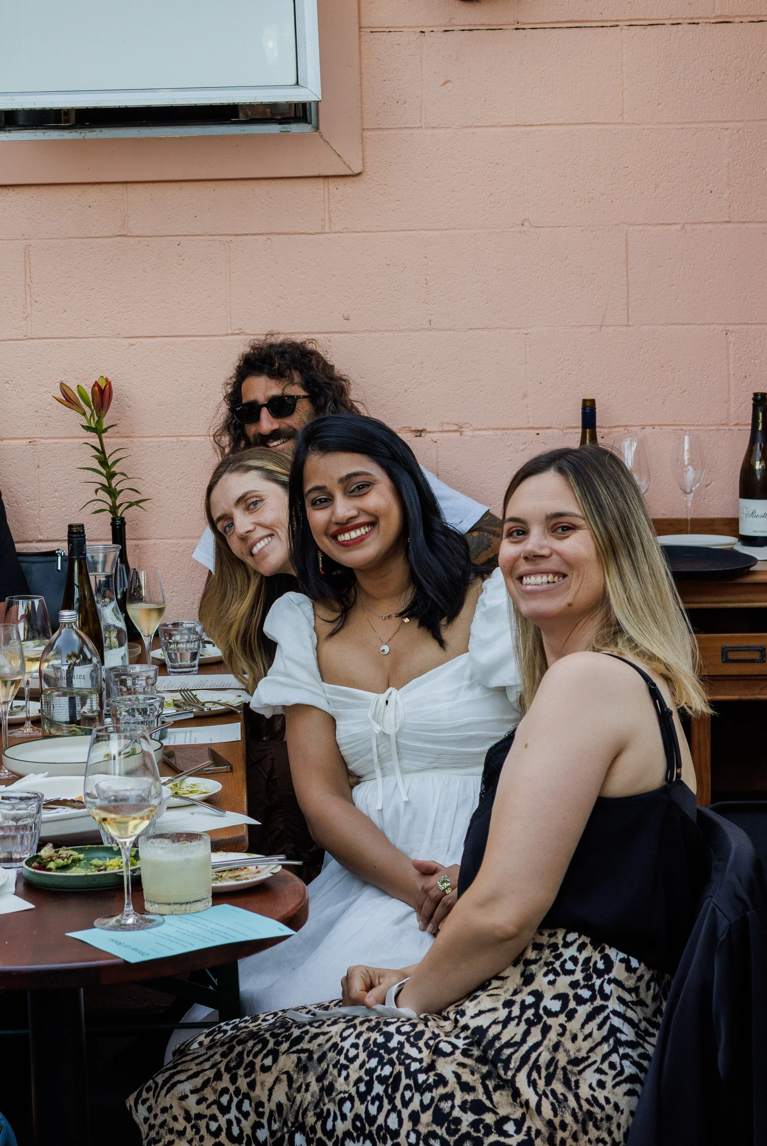 Group of four women at a dinner table smiling, with wine glasses, bottles, plates of food, and flowers in a restaurant with a peach-colored brick wall.