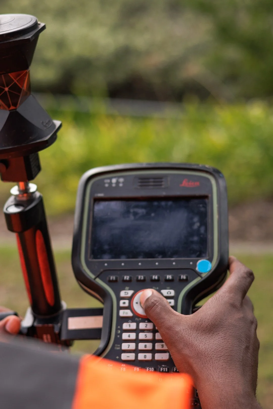 Close-up of a person's hand operating a black and gray electronic device with a screen and multiple buttons, mounted on a vehicle or equipment, with a blurred outdoor background.