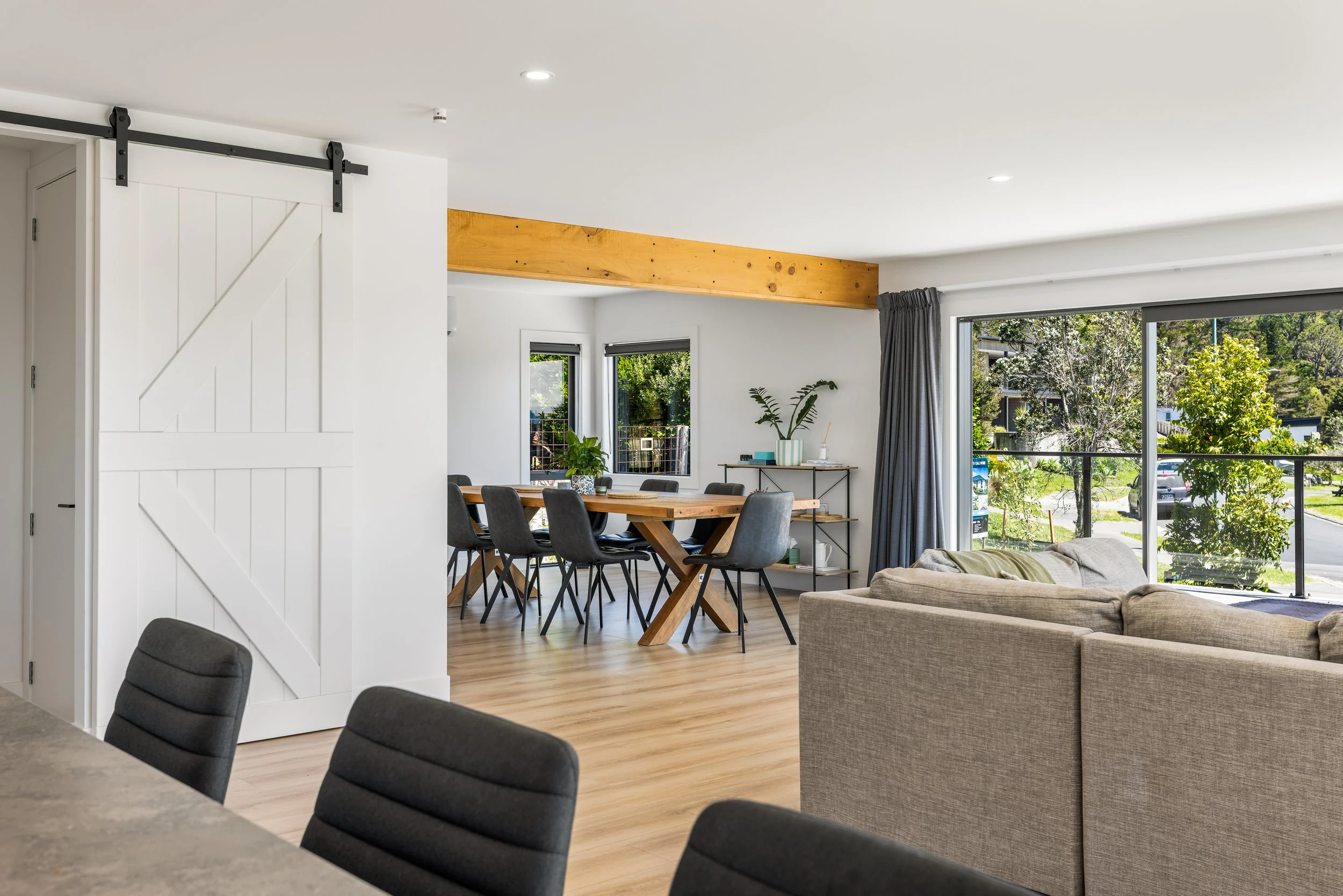 Modern living and dining room with wooden flooring, large windows with views of greenery, a dining table with black chairs, a beige sofa, and decorative plants.