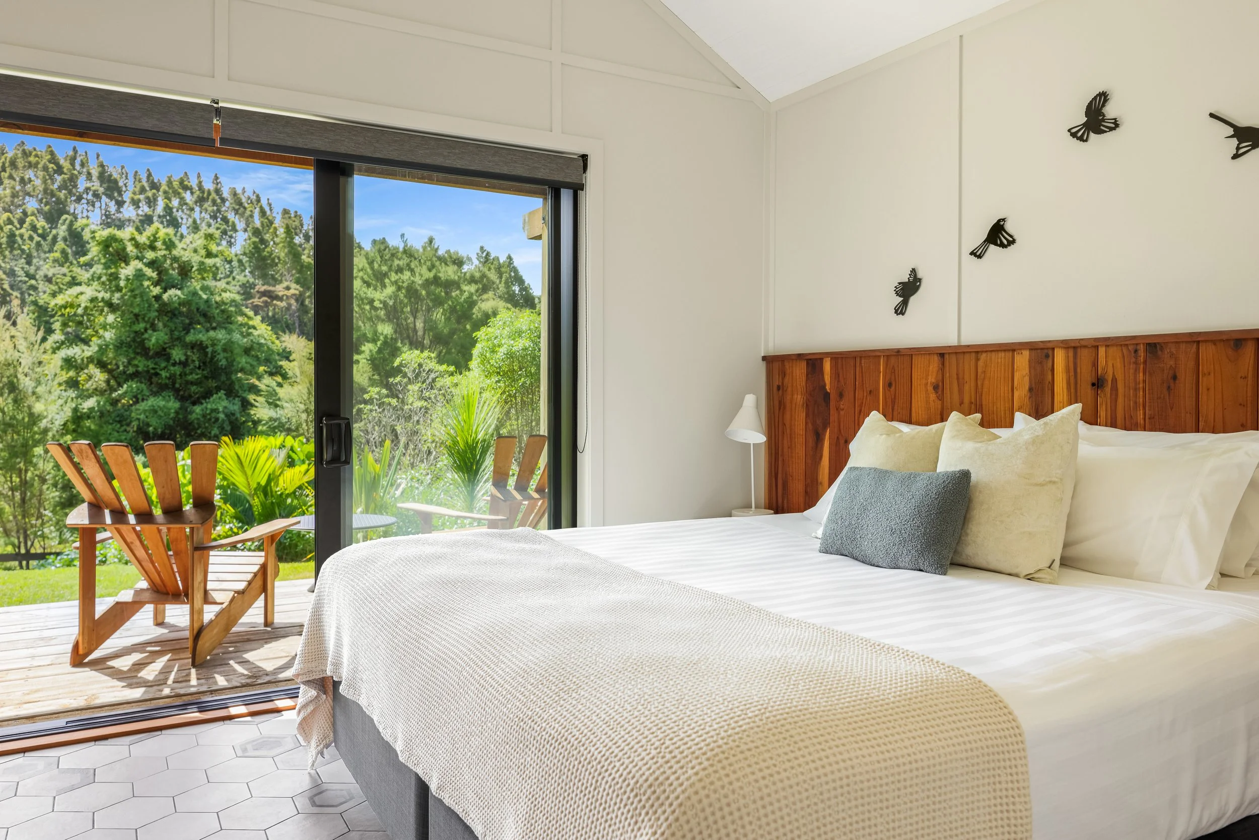 A bedroom with a bed, decorative pillows, a white lamp, and large sliding glass doors leading to a patio with outdoor chairs, overlooking green trees and a clear blue sky.