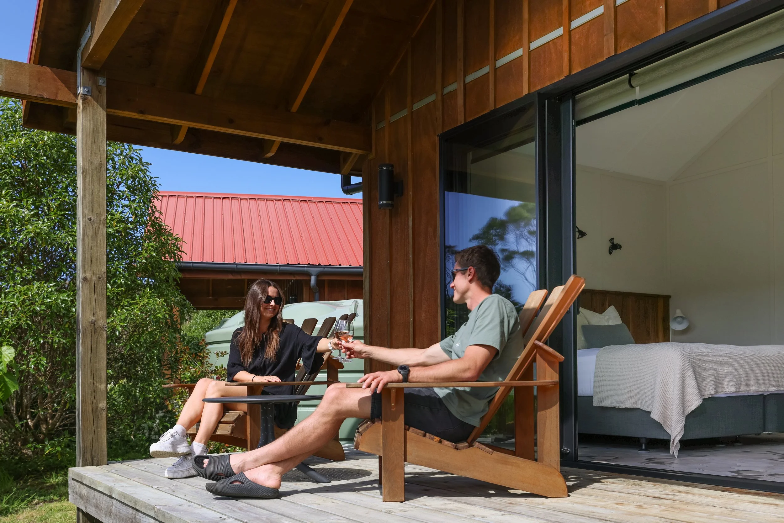 A man and woman sit on a wooden deck outside a modern house with large glass sliding doors, sharing a toast with glasses of rosé wine, surrounded by greenery and a red roof in the background.