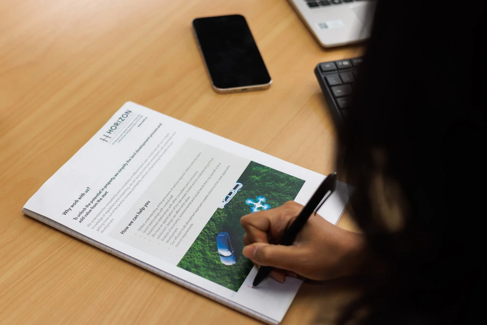 Person writing on a printed document on a wooden desk, with a smartphone, a laptop, and a keyboard nearby.