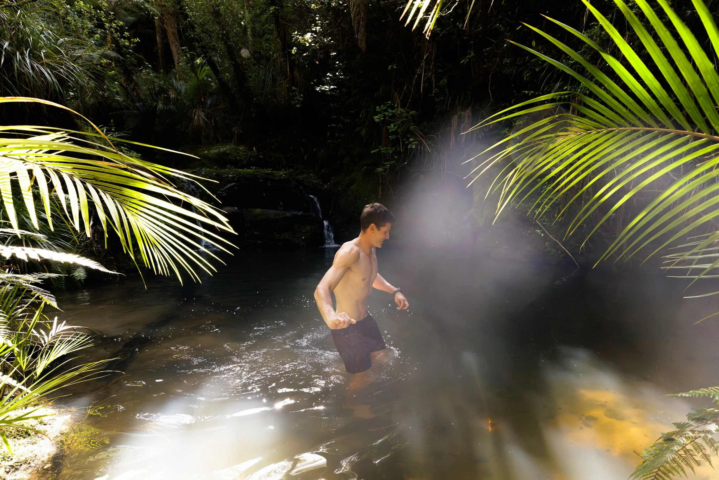 A shirtless young man is wading in a dark natural water body, surrounded by dense green tropical plants and large palm leaves, with mist rising near the water's surface.