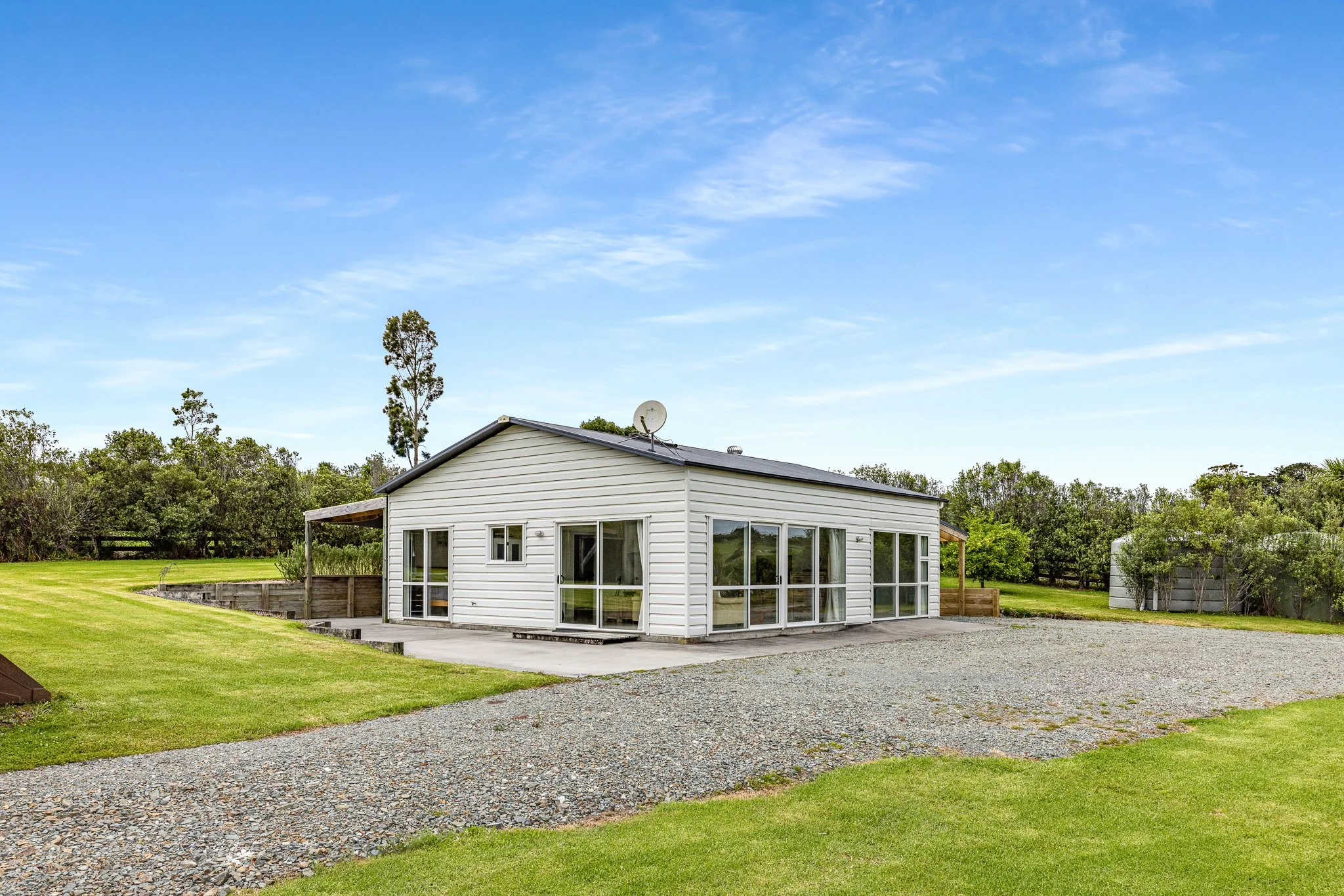 A modern white house with large glass windows and a satellite dish on the roof, set in a grassy yard with trees and a gravel driveway under a blue sky with scattered clouds.