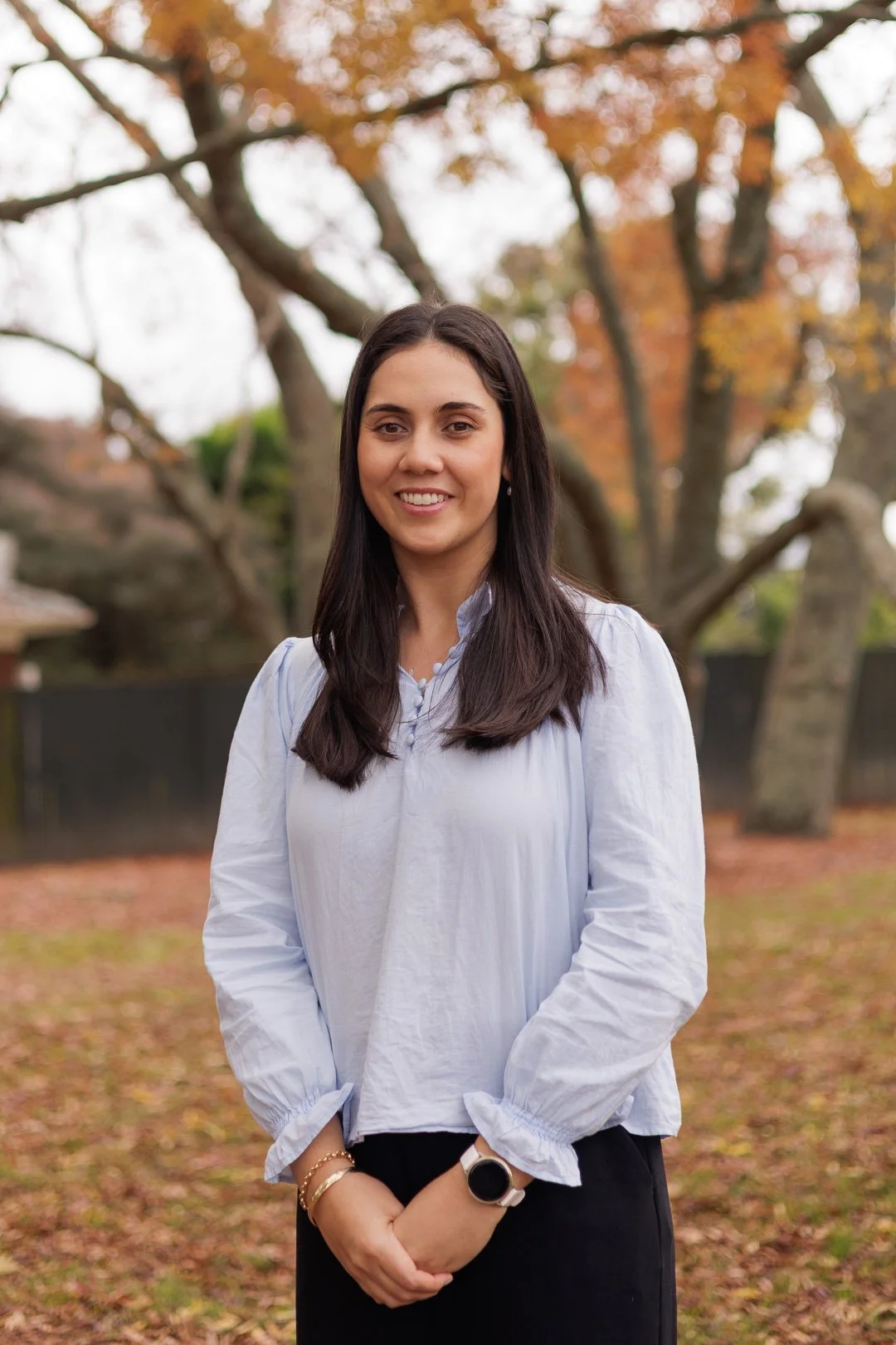 A young woman with long dark hair, wearing a white blouse and black skirt, standing outdoors on an autumn day. She is smiling with trees and fallen leaves in the background.