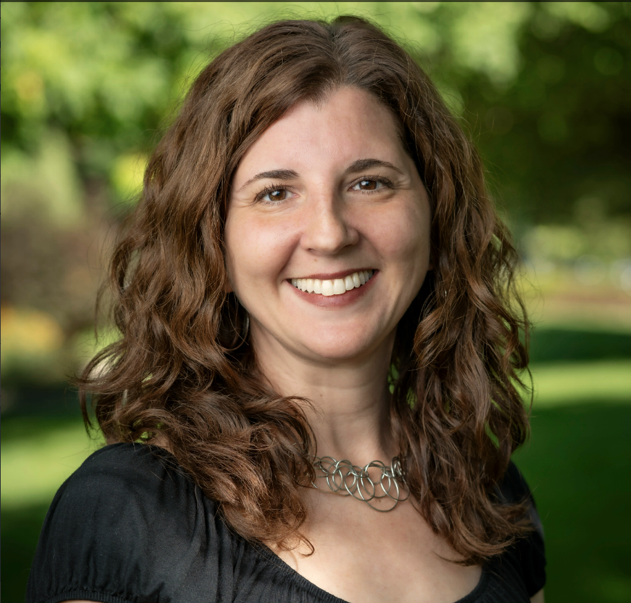 A woman with shoulder-length, curly brown hair, smiling at the camera, wearing a black top and a silver necklace, with a blurred green outdoor background.