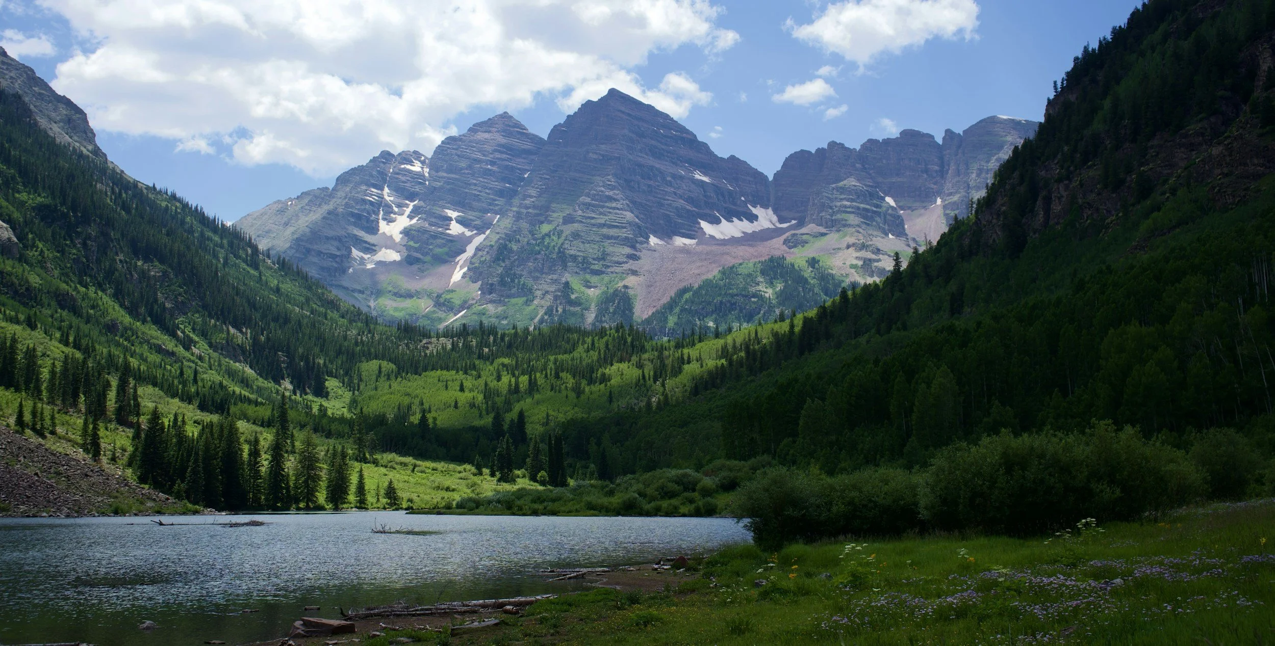 A scenic landscape featuring a lake in the foreground, surrounded by dense green forest, with towering mountains in the background under a partly cloudy sky.