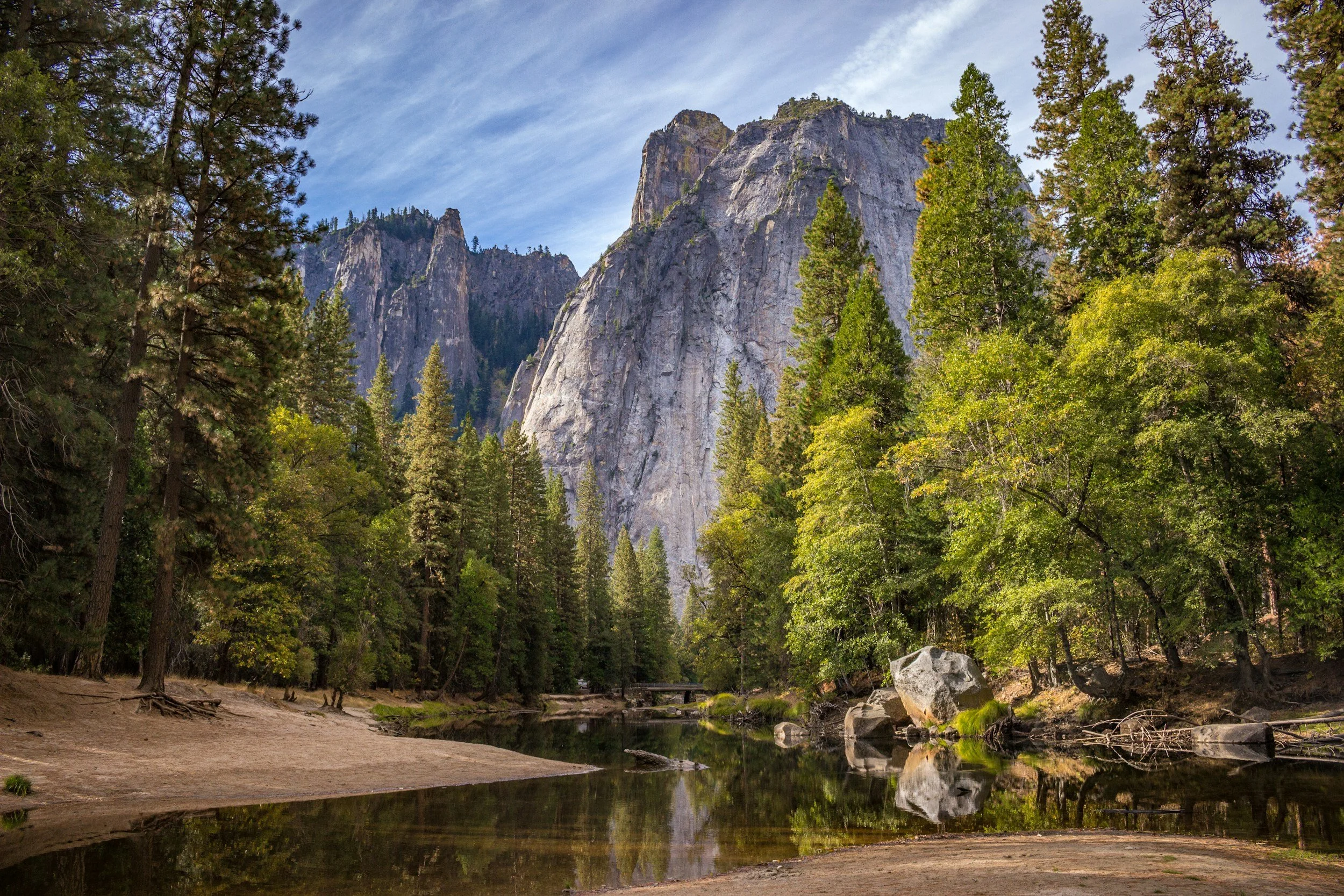 Scenic view of a forested area with towering granite cliffs in the background, a small river flowing through the forest, and a sandy shoreline in the foreground.
