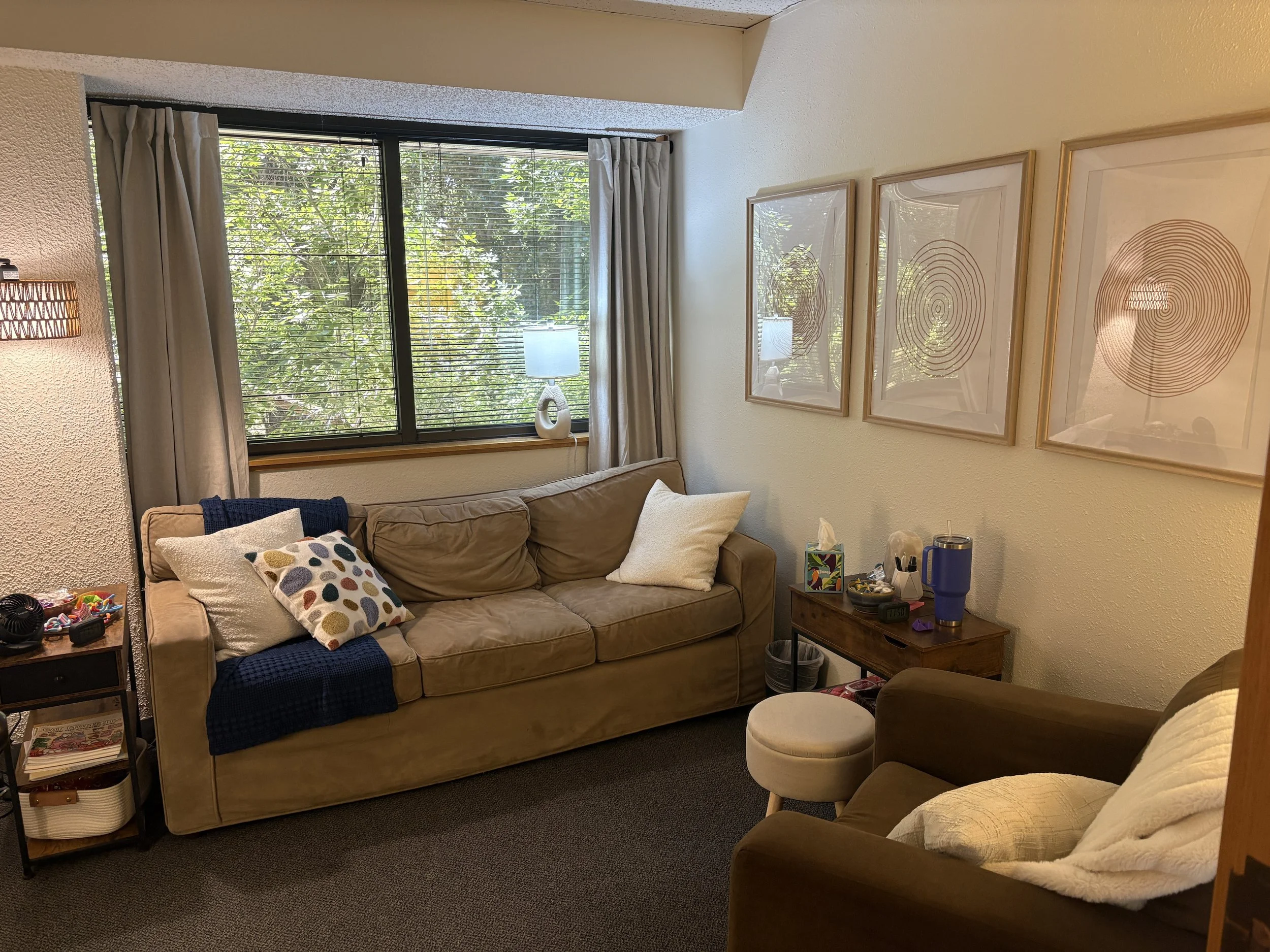 Therapy office with beige sofa, armchair, window with blinds, and artwork on the wall.