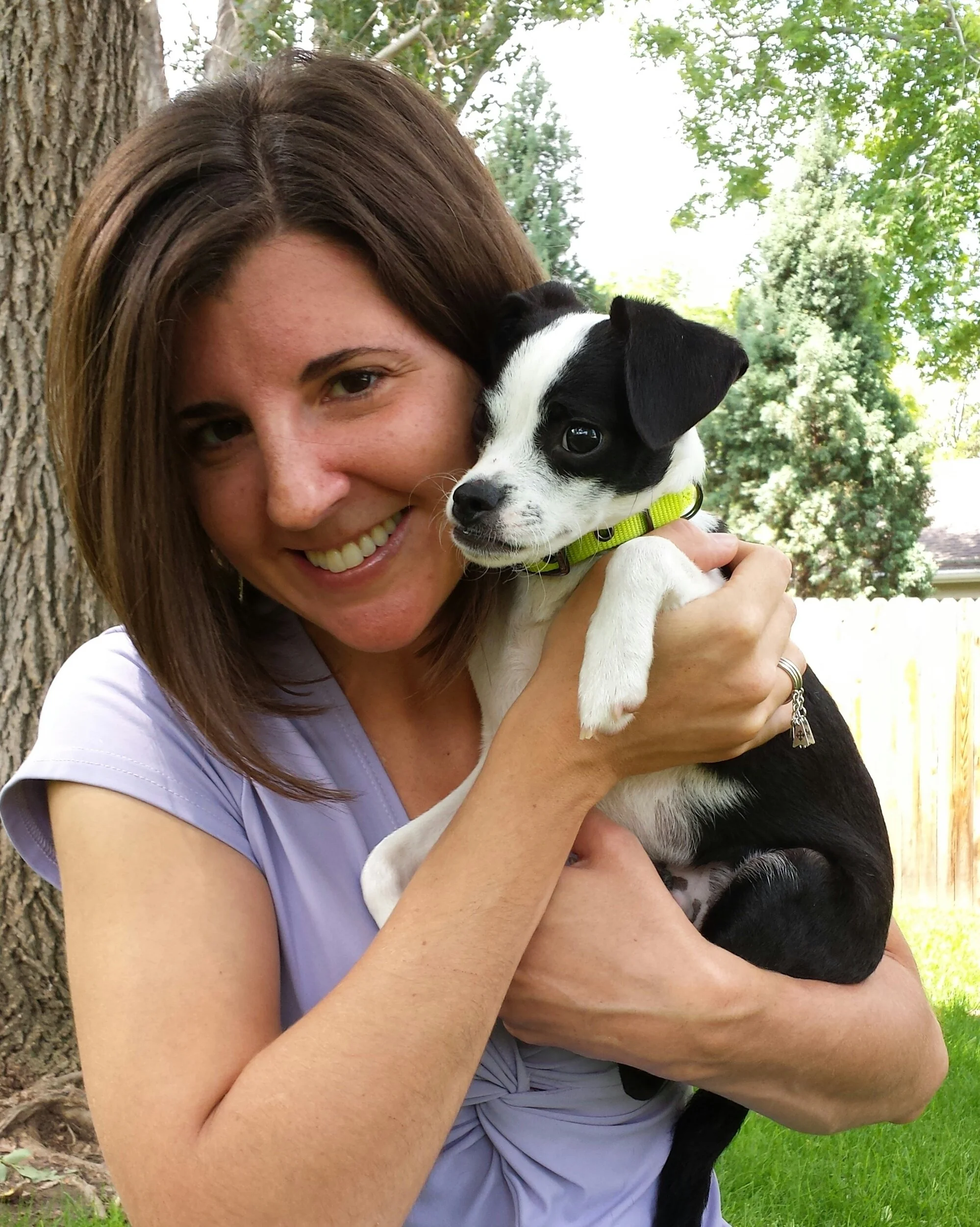 A woman smiling and holding a black and white puppy outdoors near a tree with green leaves.