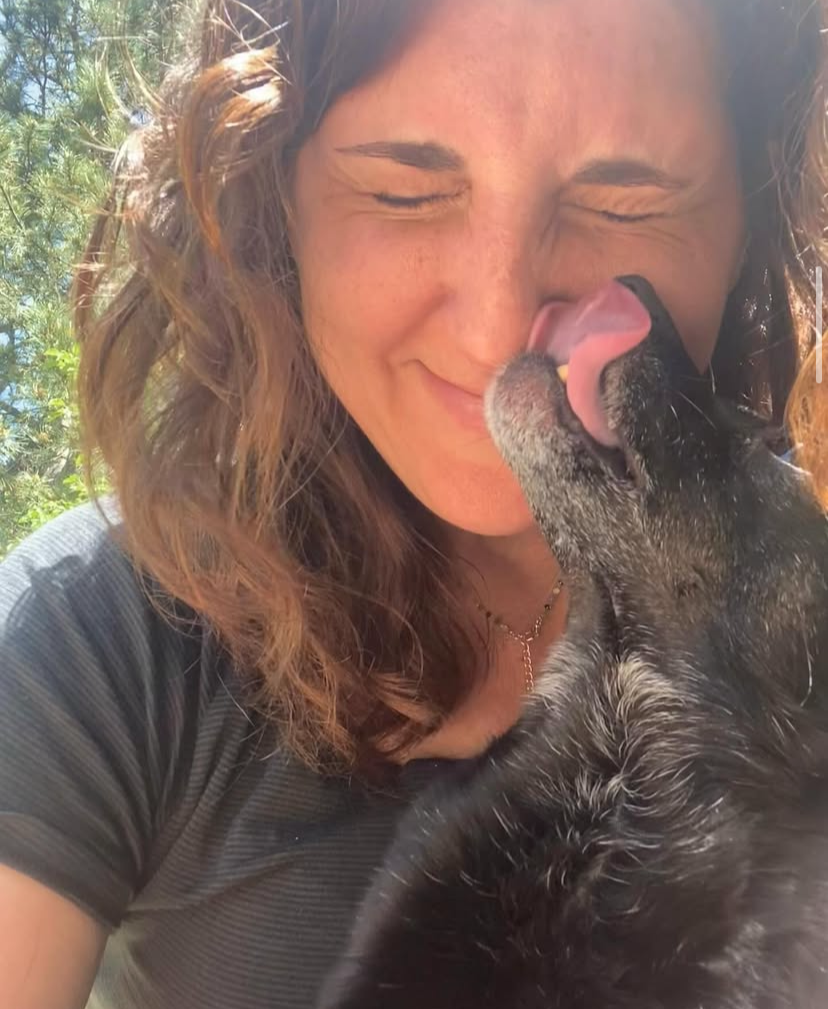A woman with curly hair and a black shirt smiling with her eyes closed, while a dog licks her outdoors with trees in the background.