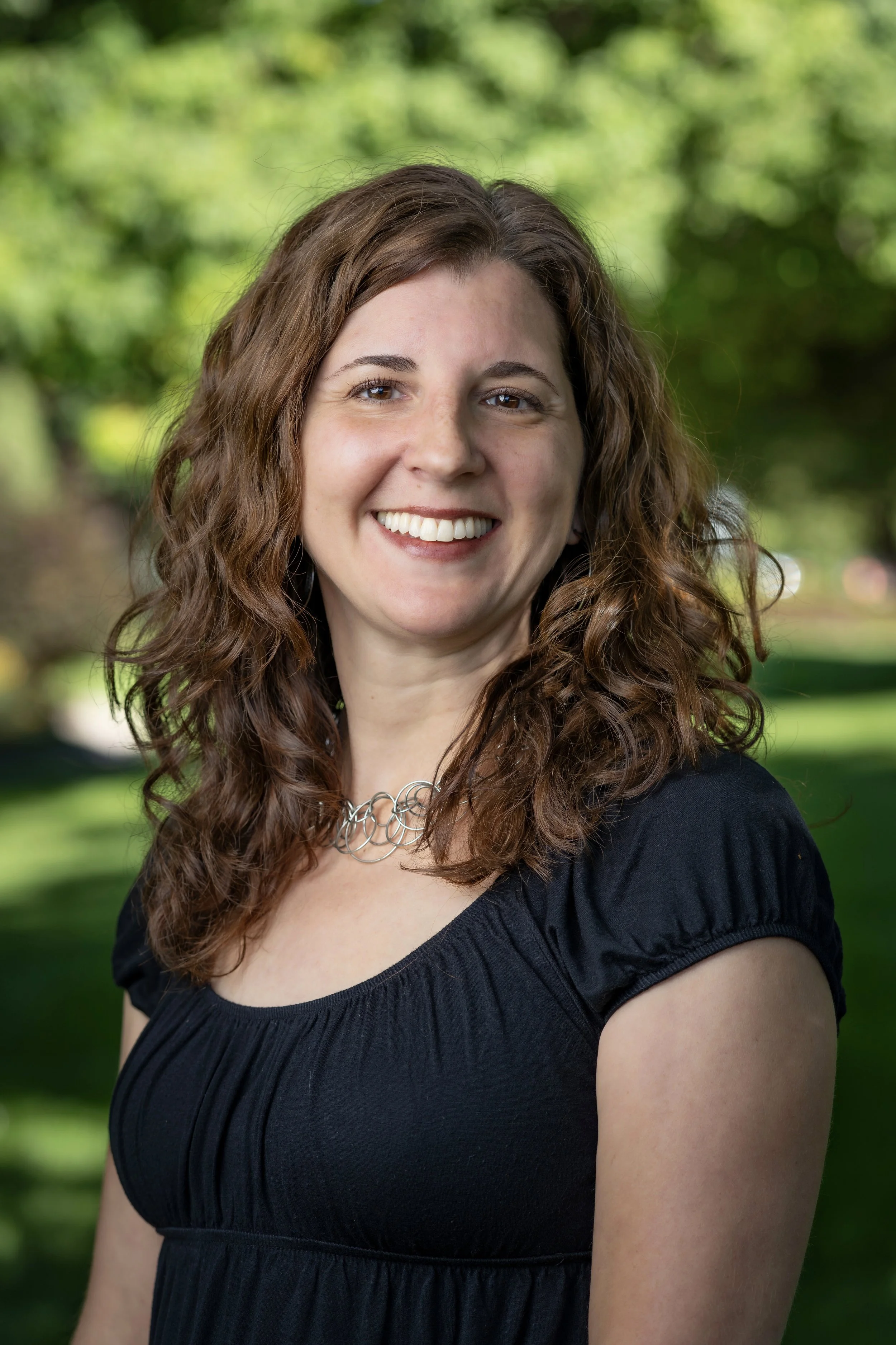 A woman with brown curly hair smiling outdoors, wearing a black shirt and a silver necklace, with a background of green trees.
