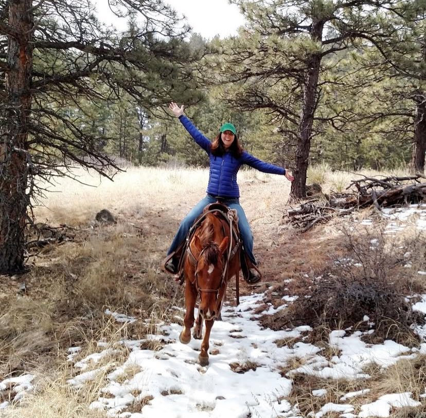 A woman wearing a green hat and a blue puffy jacket riding a brown horse on a snowy trail through a forest with pine trees, smiling with arms outstretched.