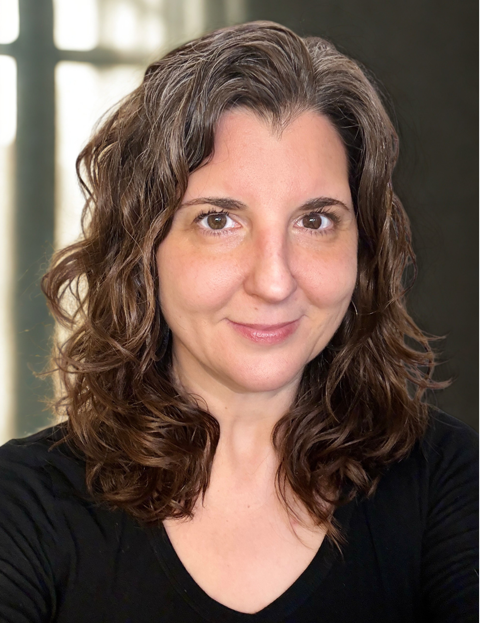 Close-up of a woman with curly brown hair and brown eyes wearing a black top, smiling softly.