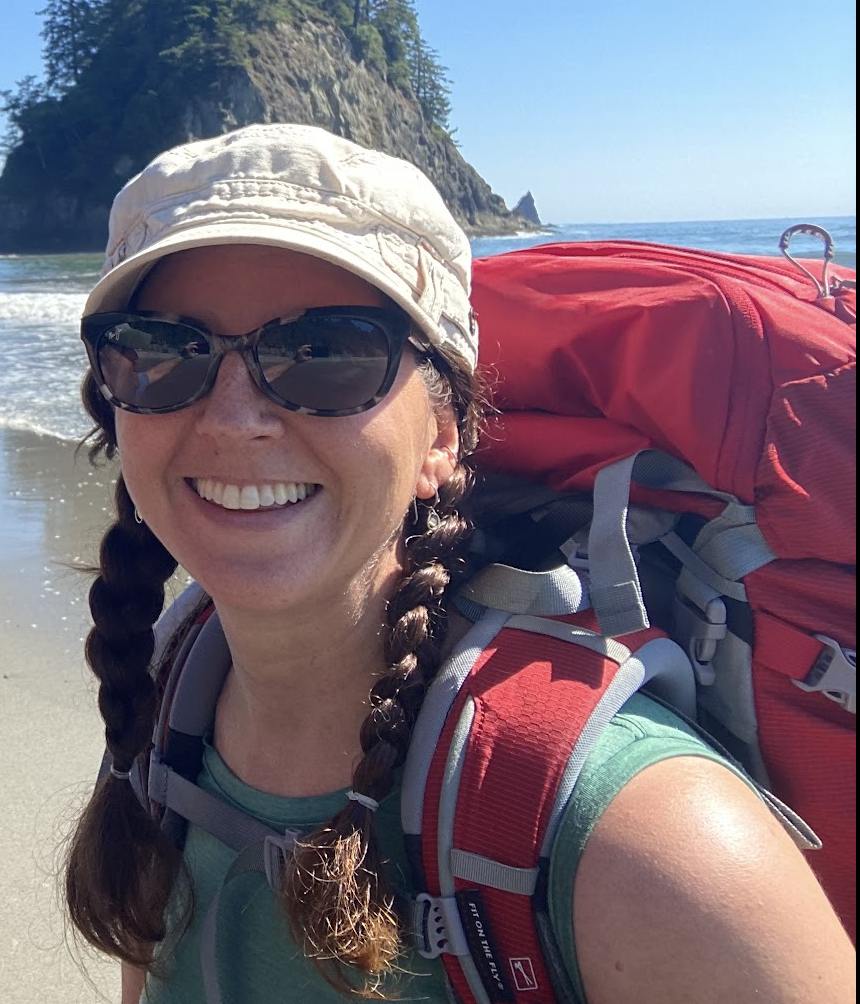 Woman smiling at the beach, wearing sunglasses, a tan cap, and a red backpack, with ocean and cliffs in the background.