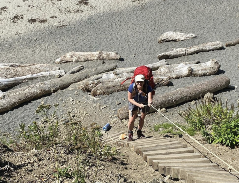 A woman backpacking up a wooden staircase on a sandy trail with large driftwood logs in the background, carrying a red pack and wearing outdoor gear.