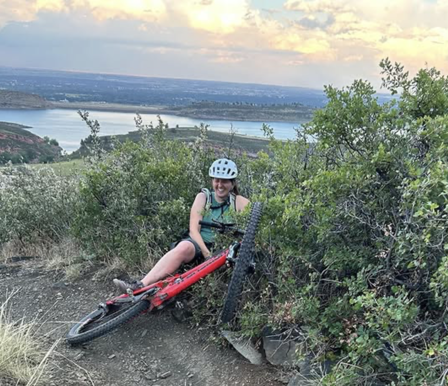 A woman wearing a helmet and a teal top sitting next to her red mountain bike on a dirt trail surrounded by green bushes and vegetation, overlooking a body of water and a scenic landscape during sunset.