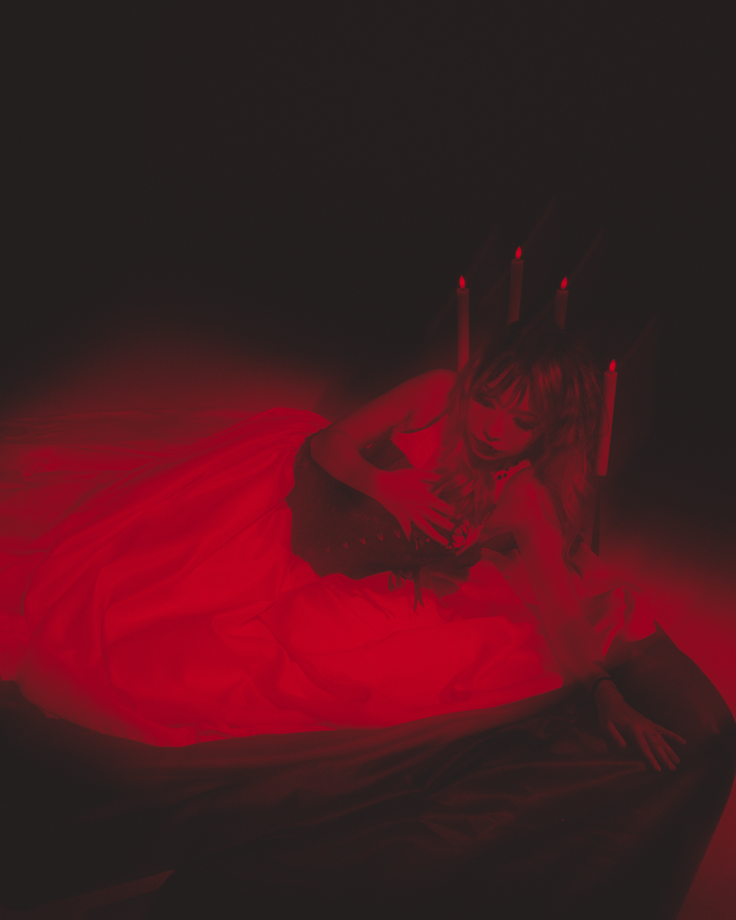 A woman with long hair lying down with her eyes closed, resting her head on her hand, dressed in a flowing red gown. The background features a dark gradient with red accents resembling flowing fabric or smoke.