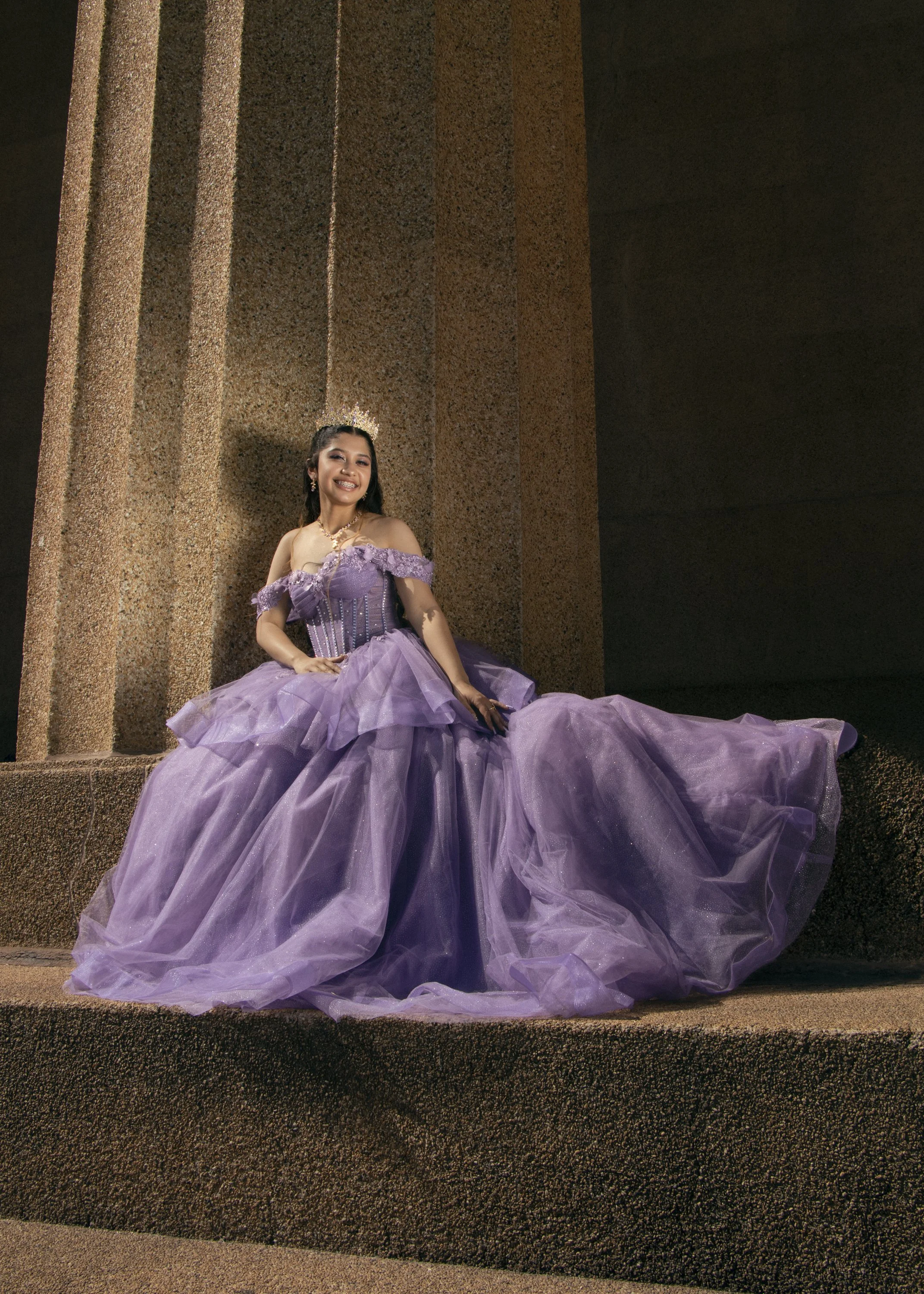 A woman dressed in a purple ball gown with off-the-shoulder sleeves, sits on stone steps in front of a textured stone wall, smiling at the camera, wearing a tiara and jewelry.