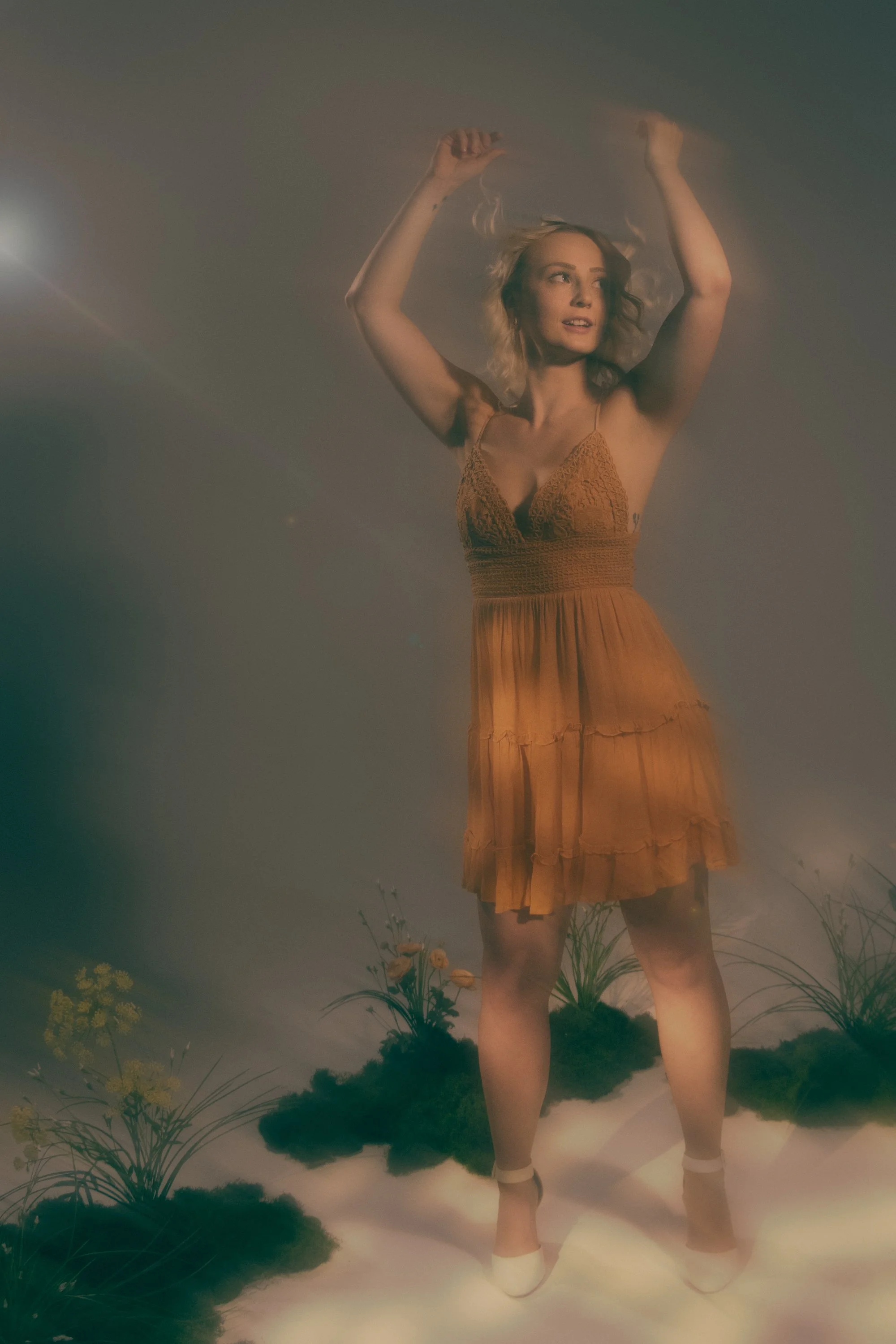 A woman in an orange dress standing among plants, with her arms raised above her head and looking to the side amidst a foggy or smoky setting.