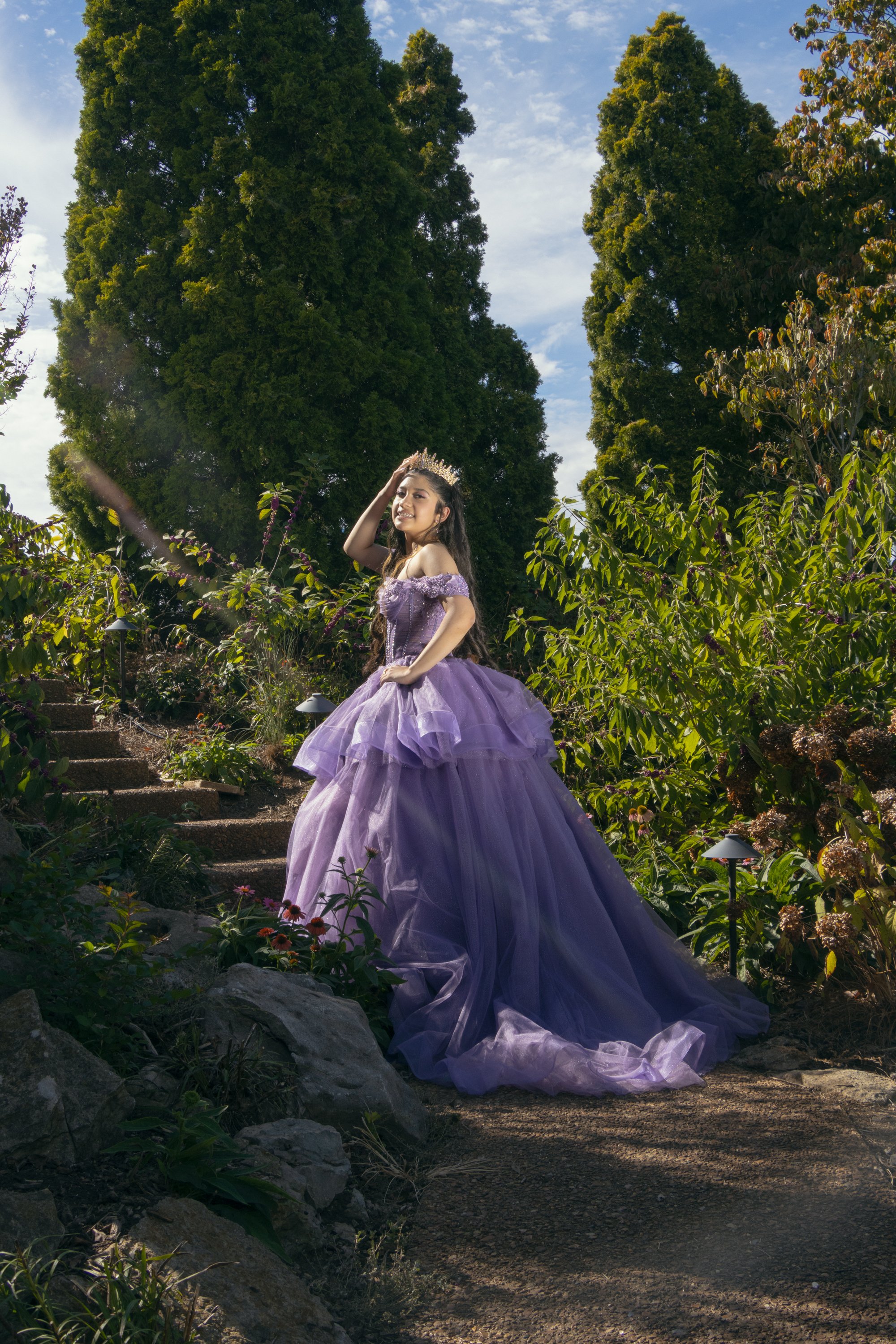 Young woman wearing a purple princess gown and a tiara, standing on a garden staircase surrounded by greenery, with large trees and a partly cloudy sky in the background.