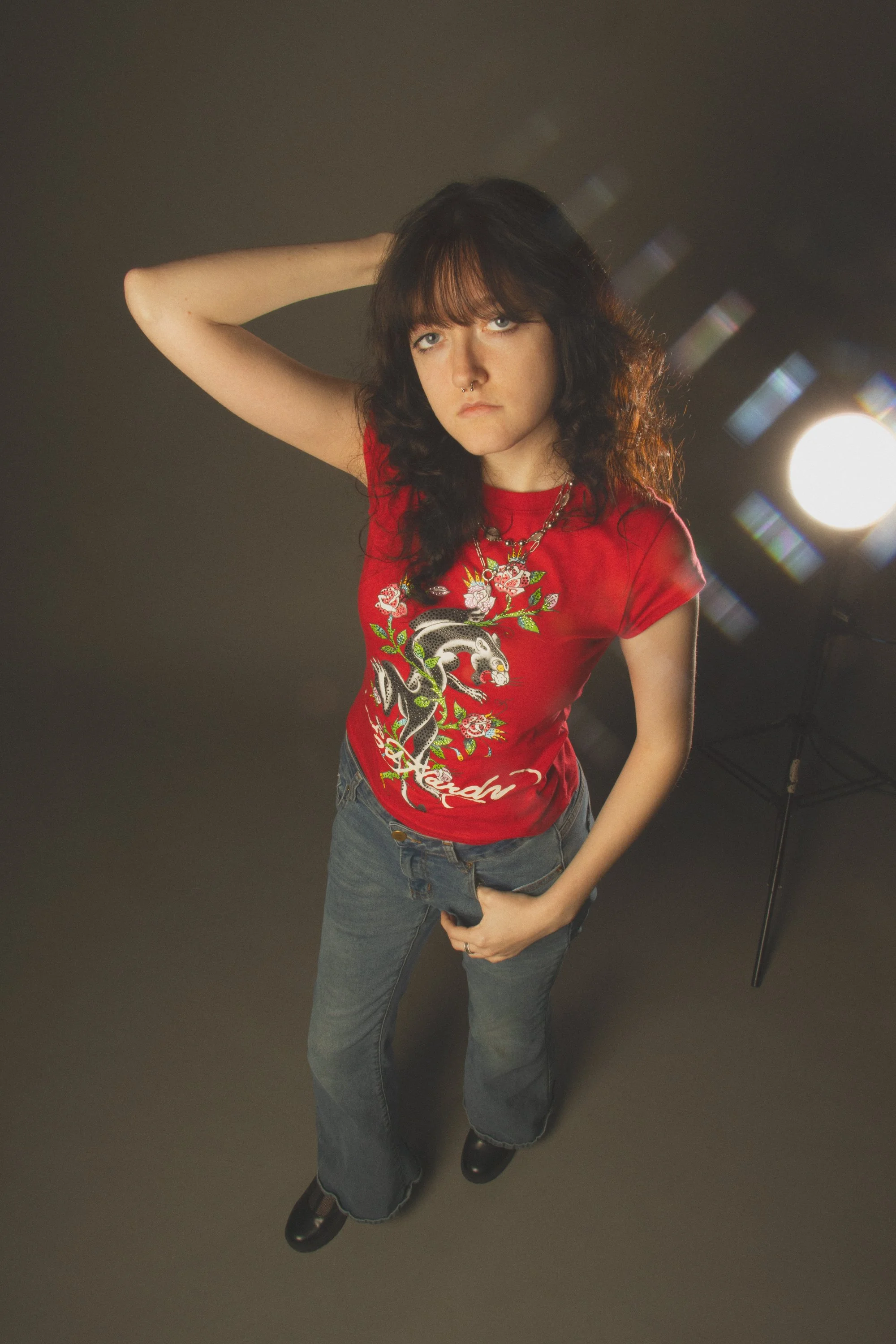 A young woman with dark wavy hair, light skin, and a serious expression, standing in front of a dark background with a bright light source behind her, wearing a red graphic t-shirt, jeans, and black shoes.