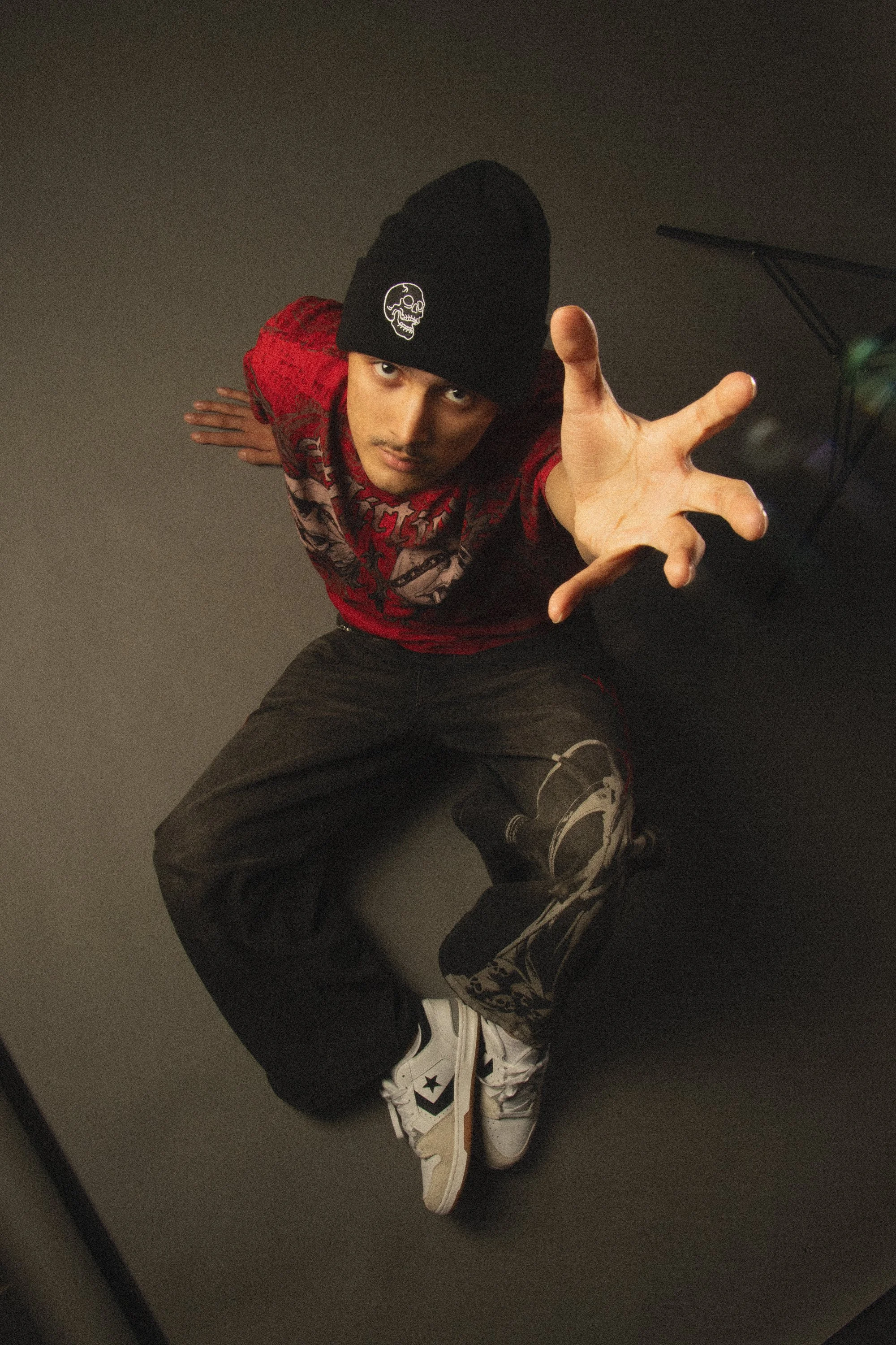A young man in a black beanie with a skull patch, red patterned shirt, black pants, and white sneakers with black stars kneeling on a gray floor reaching toward the camera.
