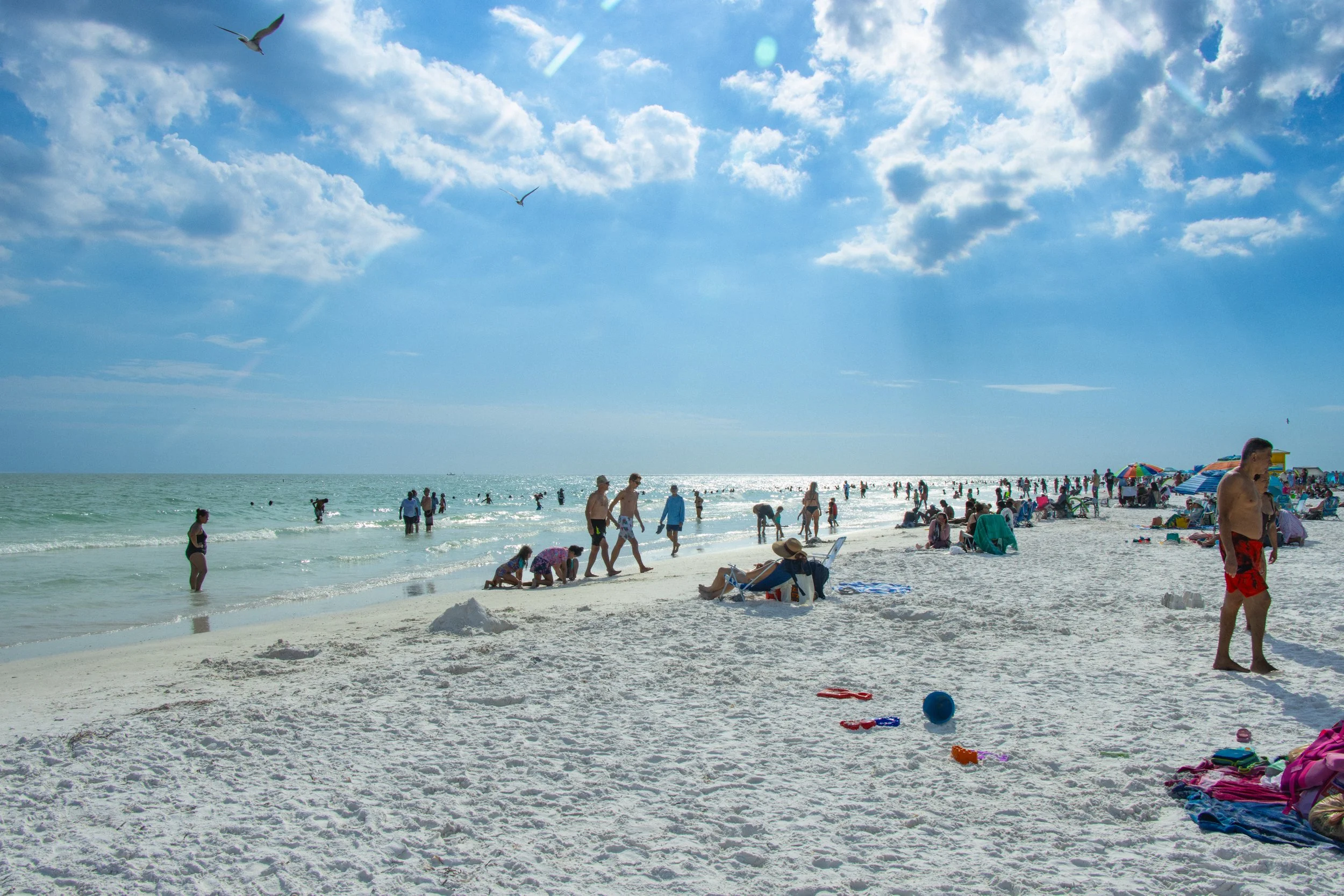 Crowded beach with people swimming, walking, and lounging on the sand under a partly cloudy sky with seagulls flying overhead.