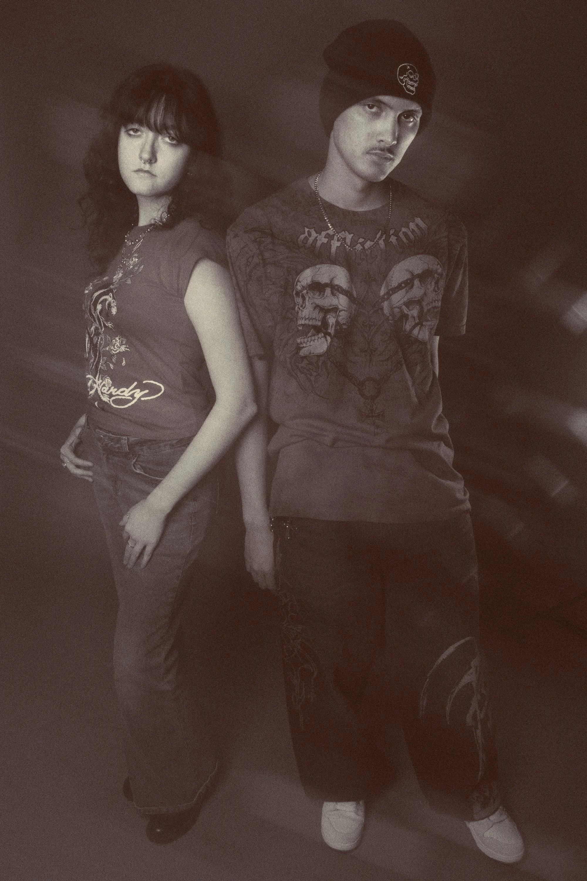 Two young individuals, a woman with wavy hair and a septum piercing, and a man with a beanie with a skull patch, standing side by side with serious expressions. The woman is wearing a graphic t-shirt with embroidered details, and the man has a t-shir