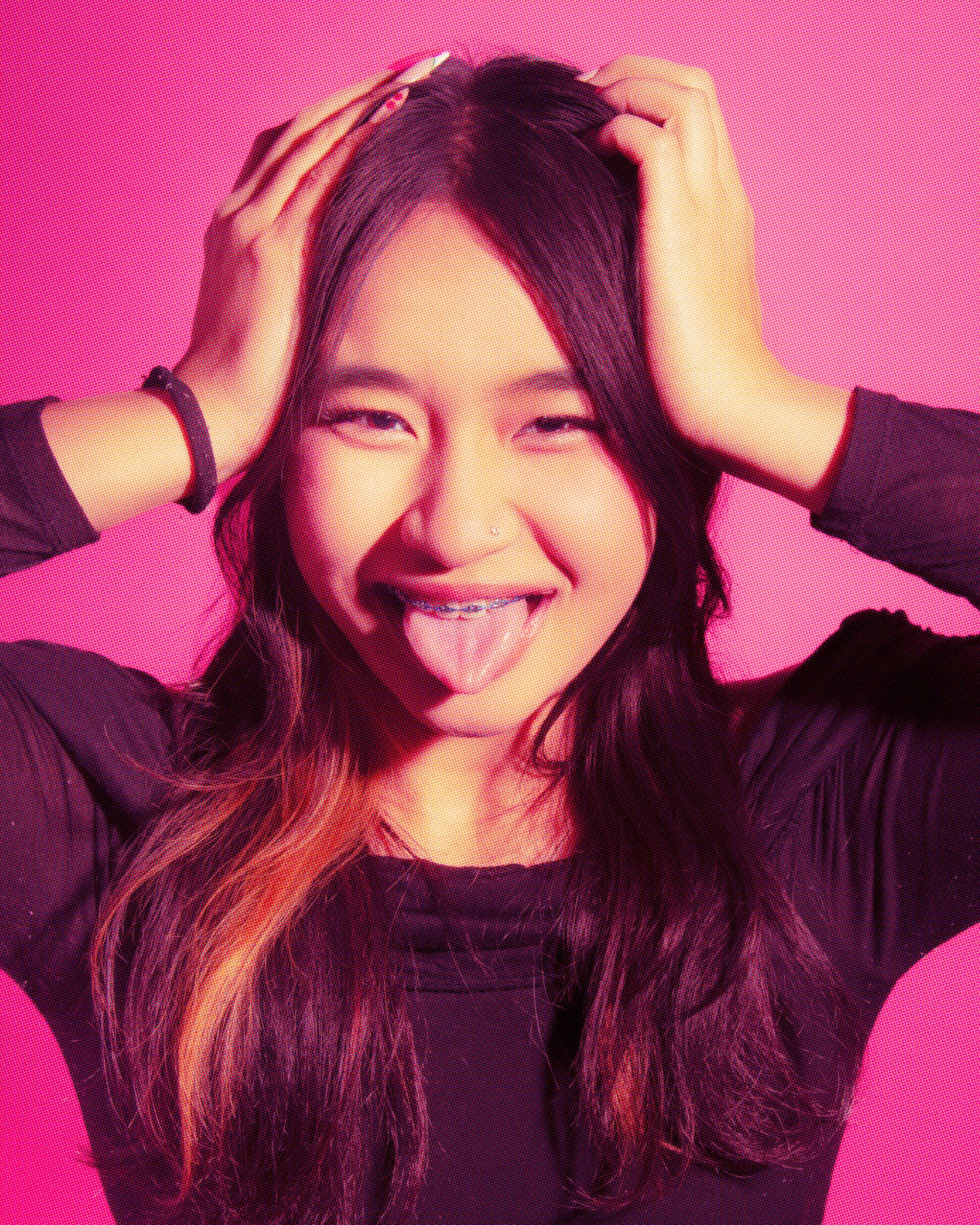 A young woman with long, dark hair, smiling and sticking out her tongue, holding her hair with both hands, against a bright pink background.