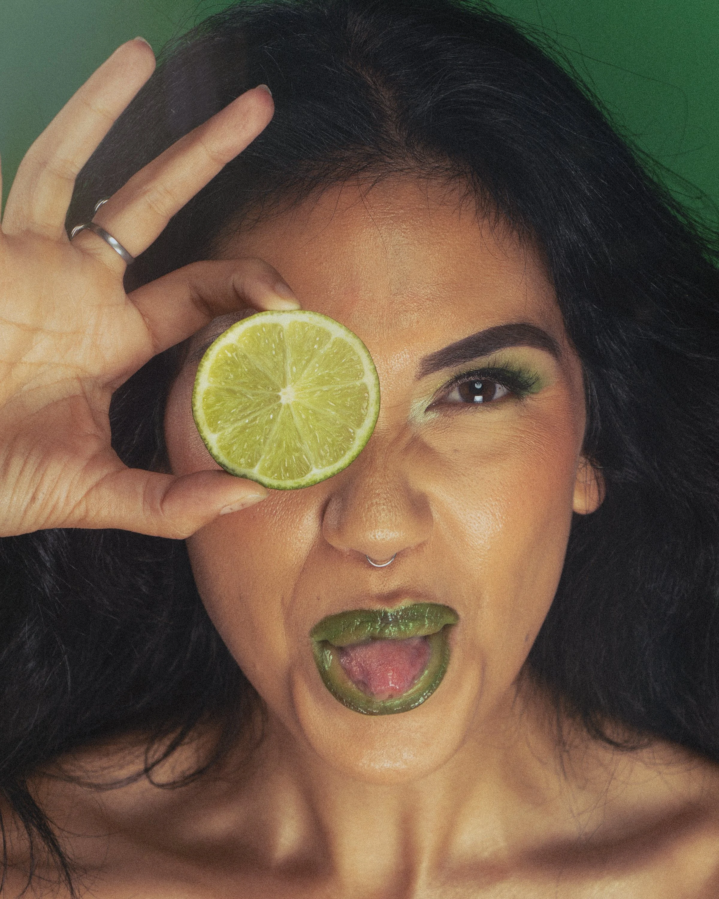 A woman with dark hair and makeup holding a lime slice over her eye, with green lipstick matching the lime, and a green background.