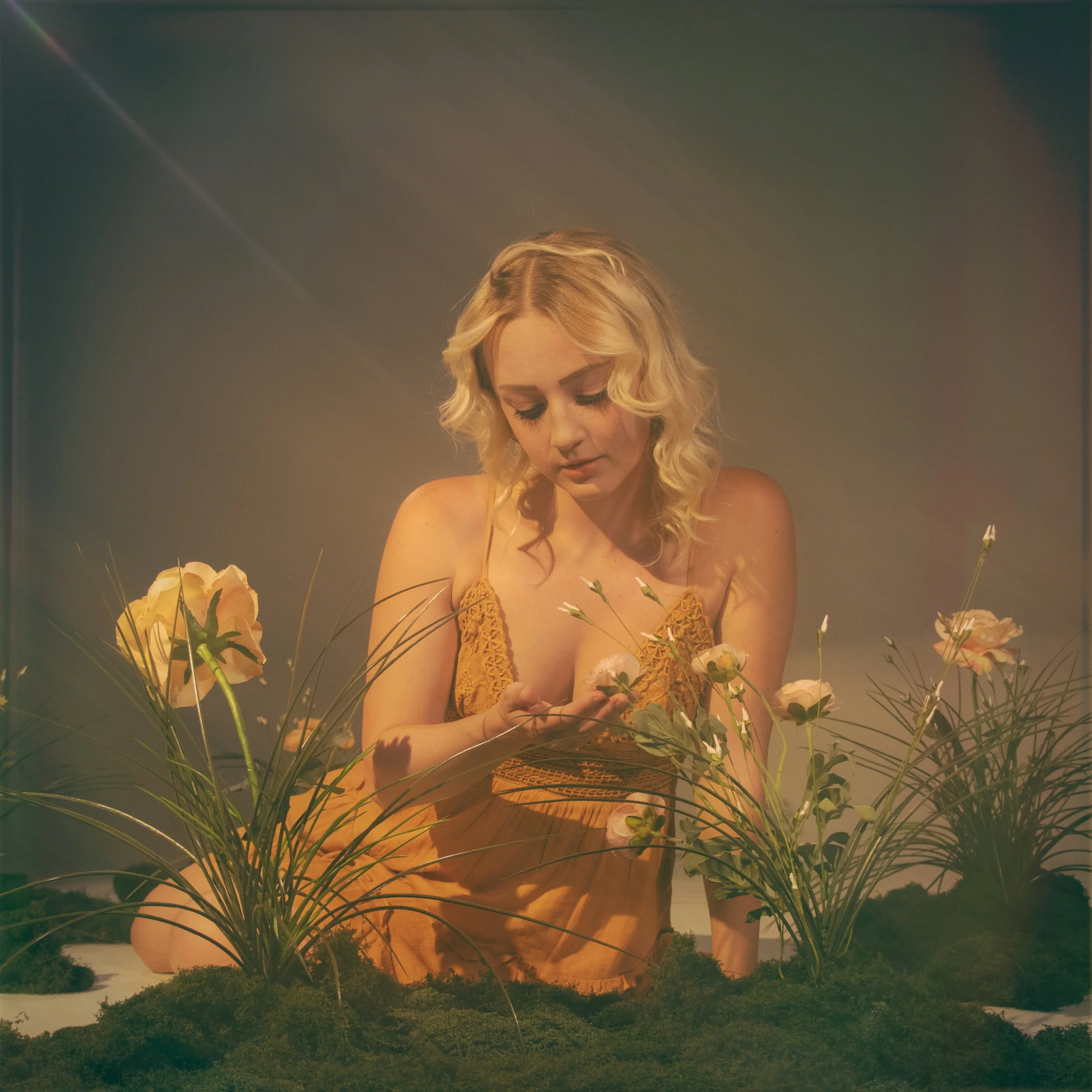 A woman with blonde curly hair wearing a yellow dress surrounded by flowers and greenery, looking down with a contemplative expression.
