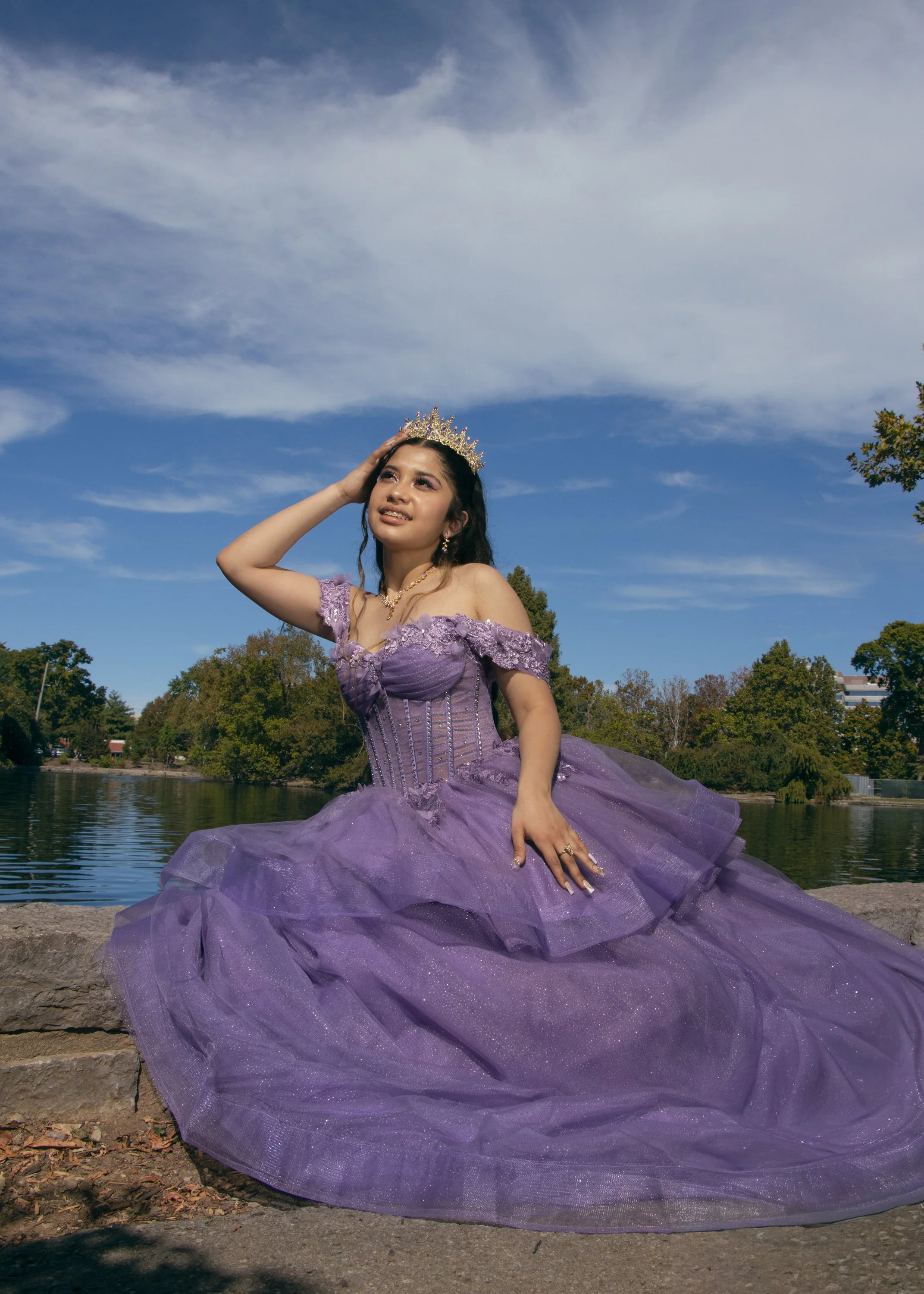 A young woman in a purple dress with lace and tulle, wearing a crown, is sitting outdoors near a body of water with trees and a partly cloudy sky in the background. She is resting her hand on her head and smiling.