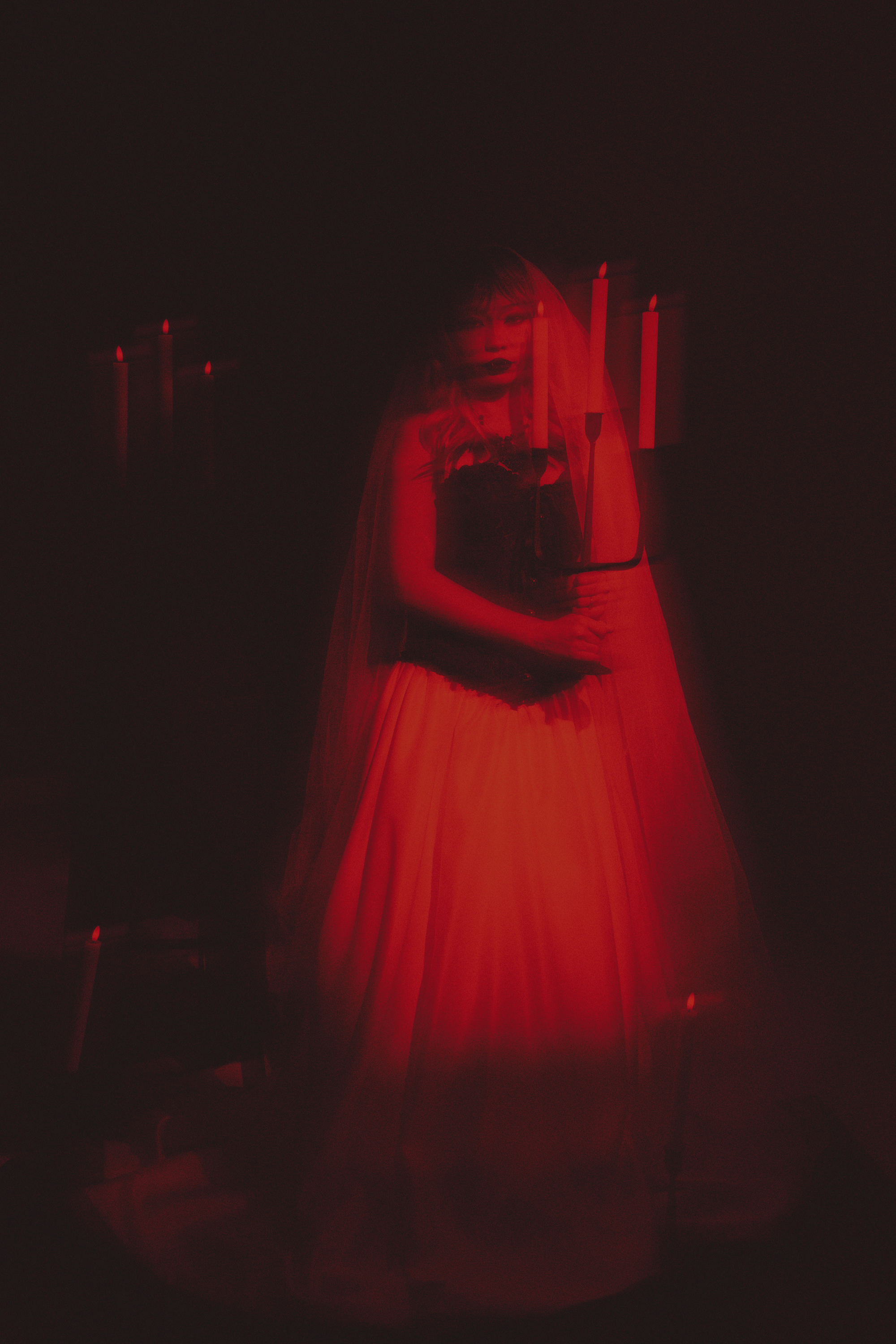 A woman in a black corset dress with a long, red, flowing skirt stands in a dark room illuminated by red candles, creating a gothic, mysterious atmosphere.