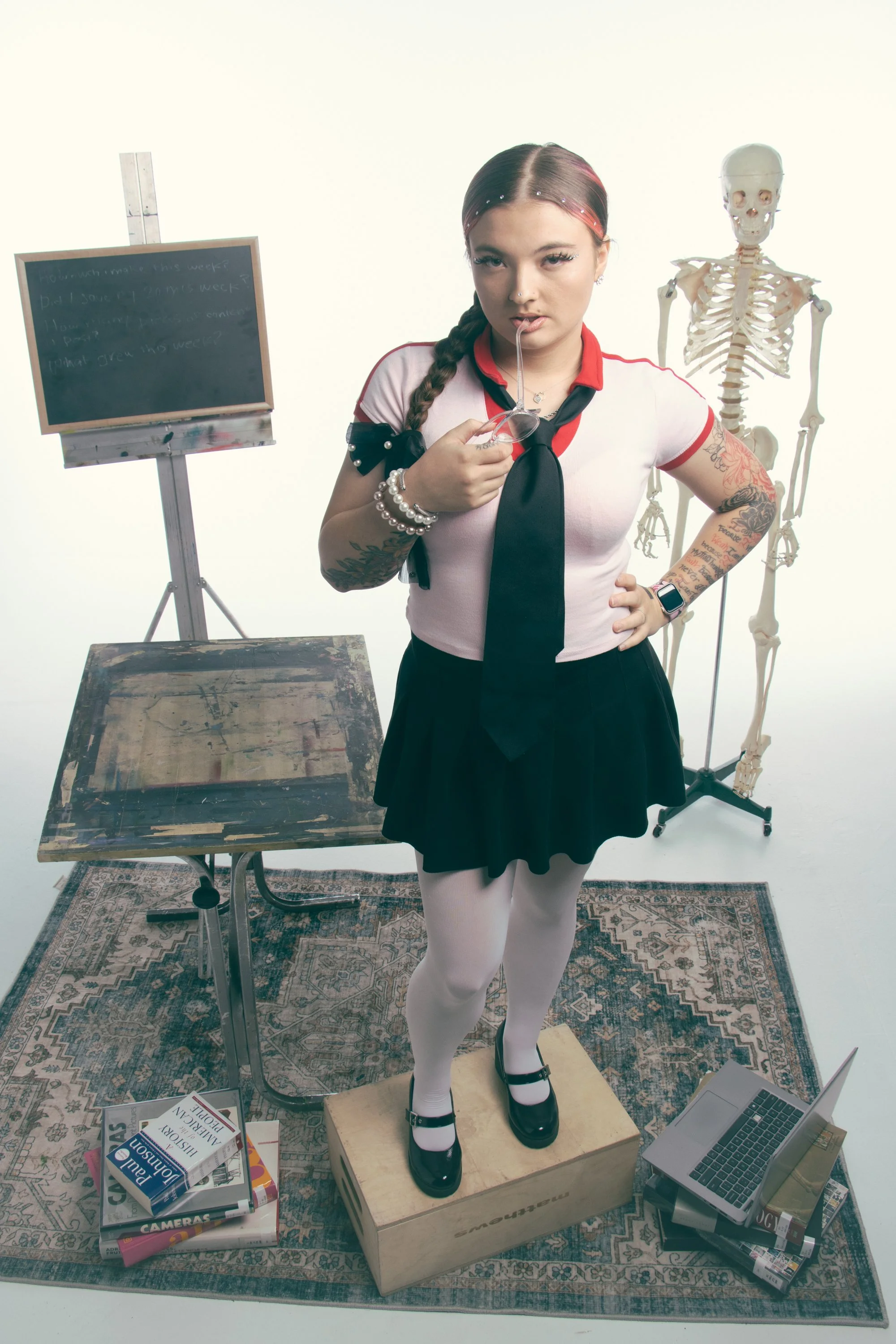 A young woman with tattoos on her arms, wearing a schoolgirl outfit, stands on a wooden box in a room with a skeleton model behind her. She is holding glasses in her mouth and has one hand on her hip. There is a blackboard on a stand, a messy desk, a