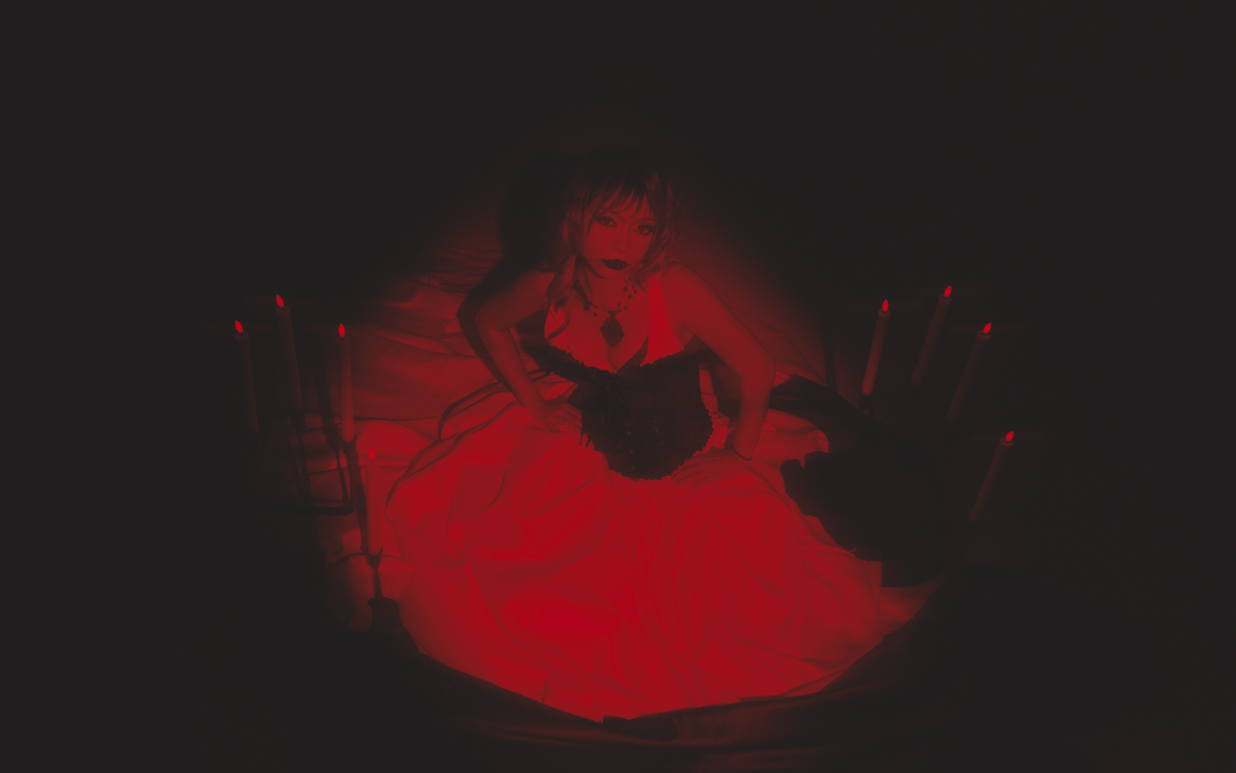 A woman with short hair and dark makeup dressed in black, lying on a bed with red lighting. She is surrounded by black candles and wearing gothic jewelry.