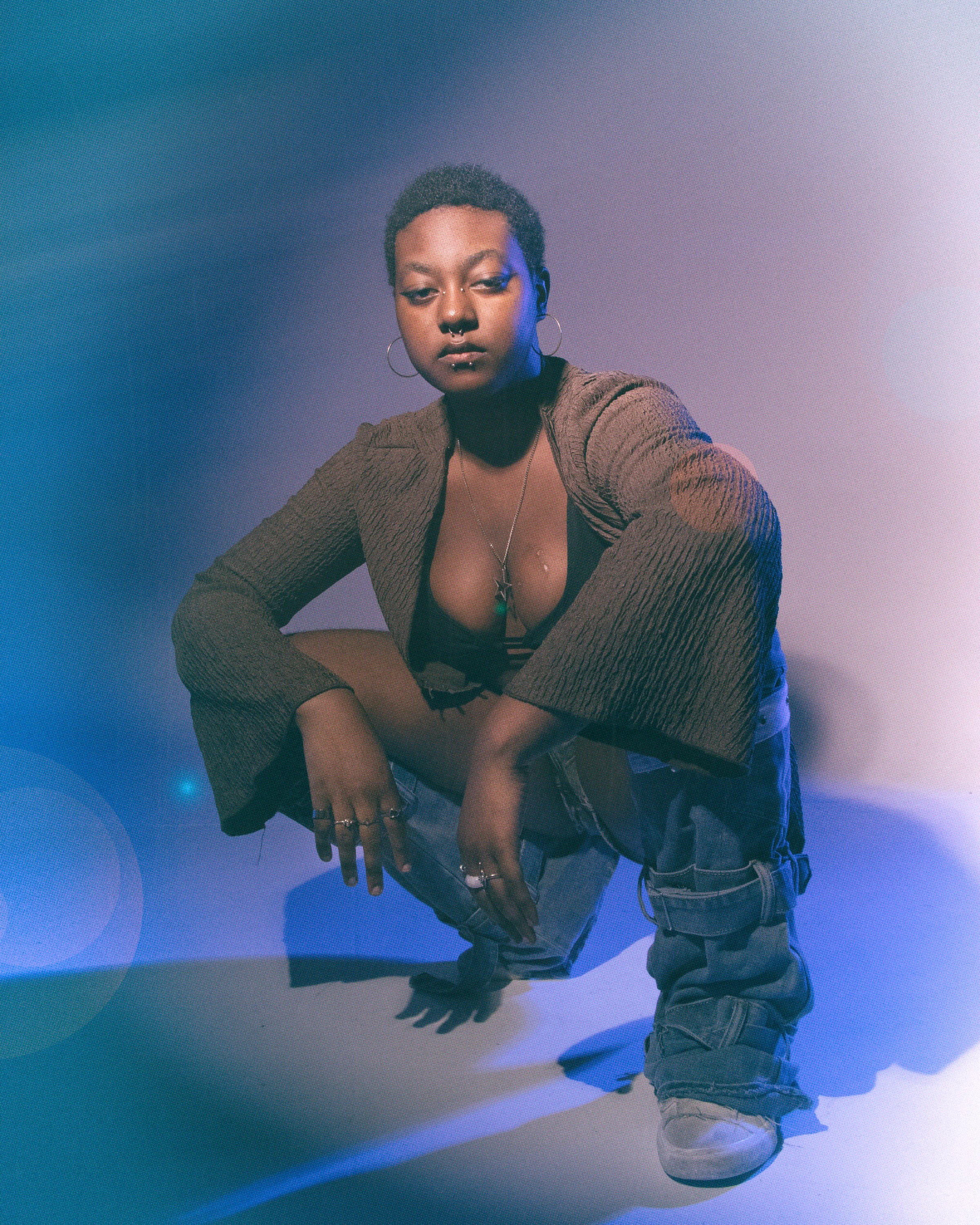 A person with short curly hair, tattoos on their arm, and piercings, wearing a brown textured blazer over a black top, and blue jeans. They are crouched down with one hand on the ground and one knee raised, with colorful lighting creating a purple an