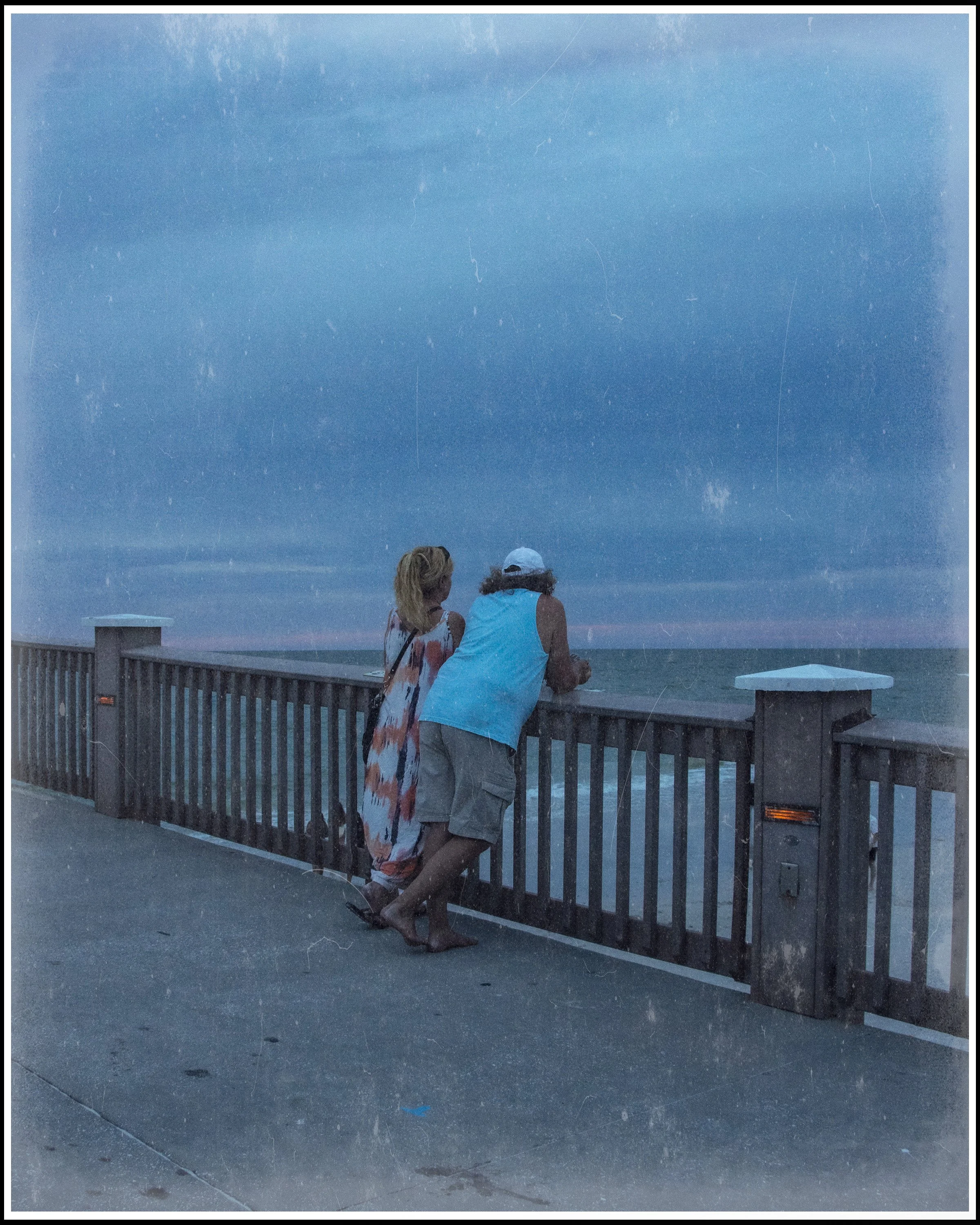 Two women leaning over a wooden railing by the beach at dusk, looking at the ocean under a starry sky.