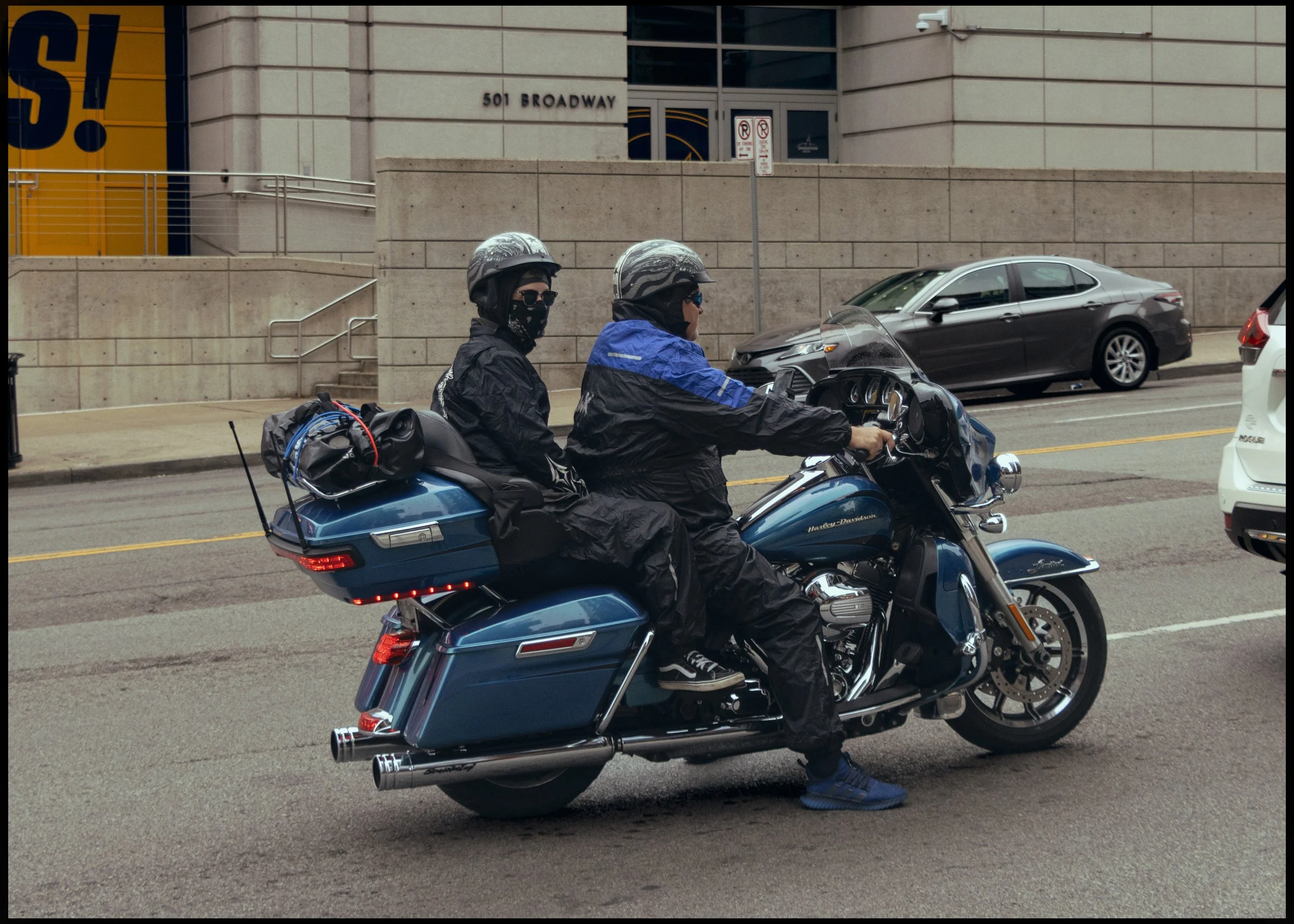Two motorcyclists wearing helmets, sunglasses, and jackets riding a blue Harley-Davidson touring motorcycle on an urban street with cars parked on the side and a large building in the background.