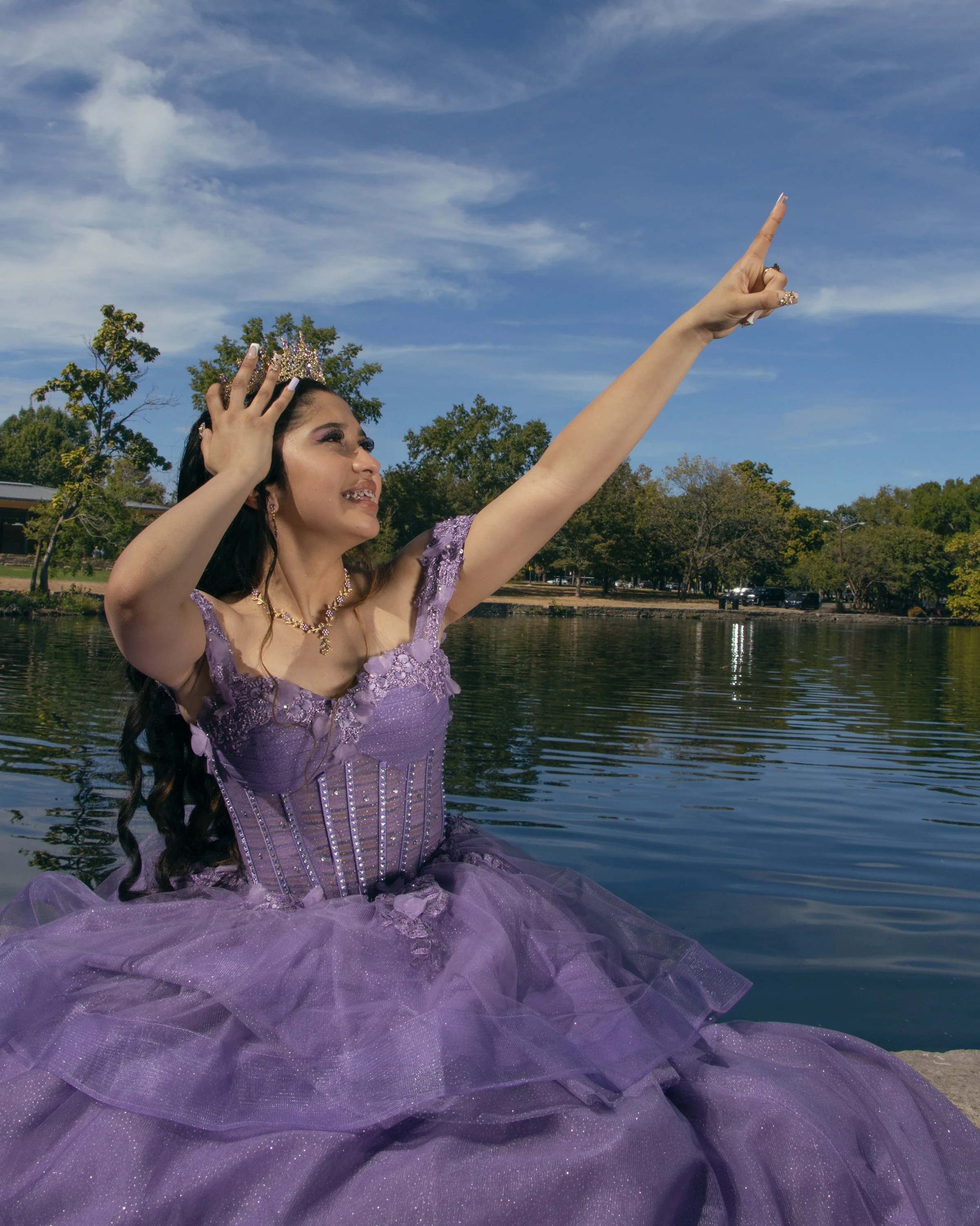 A woman wearing a purple gown and a crown, sitting in a water body surrounded by trees, with her arm extended upwards pointing at the sky.