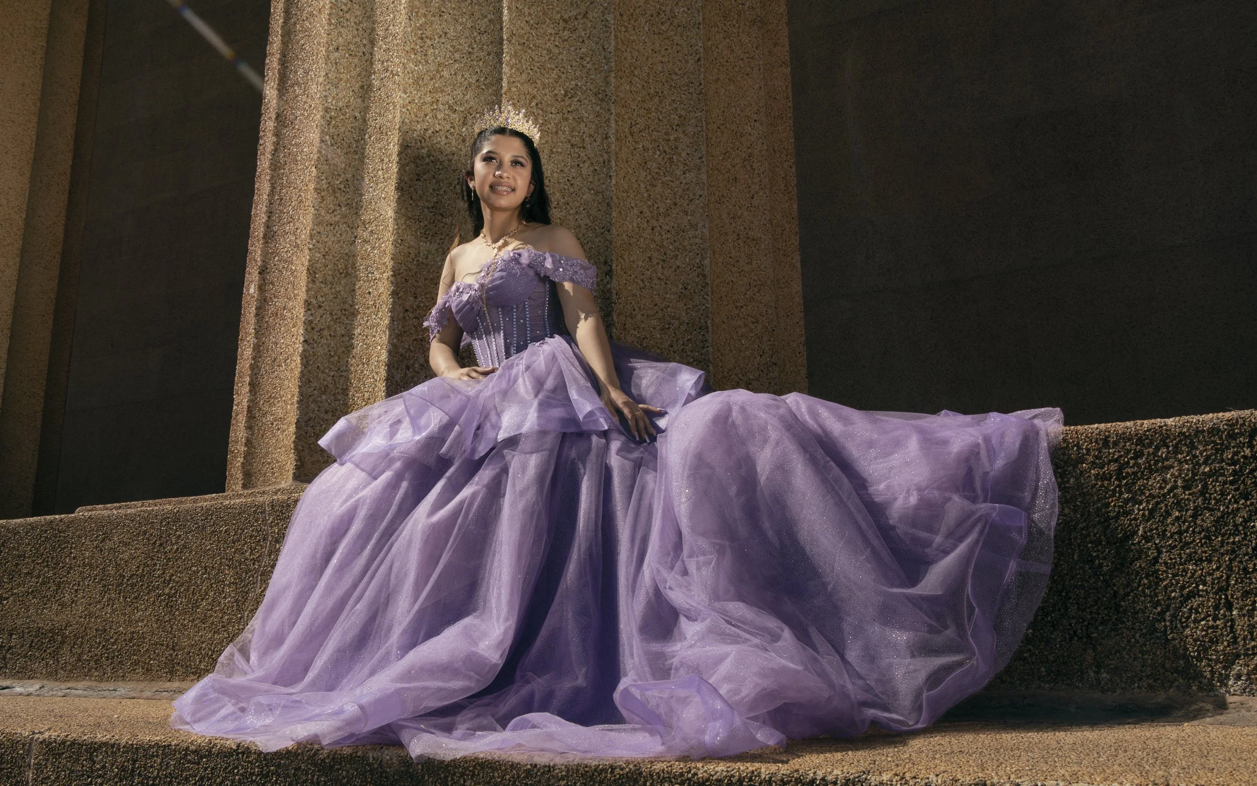 A woman in a purple ball gown sitting on stone steps outside. She wears a tiara and looks upward with a smile.