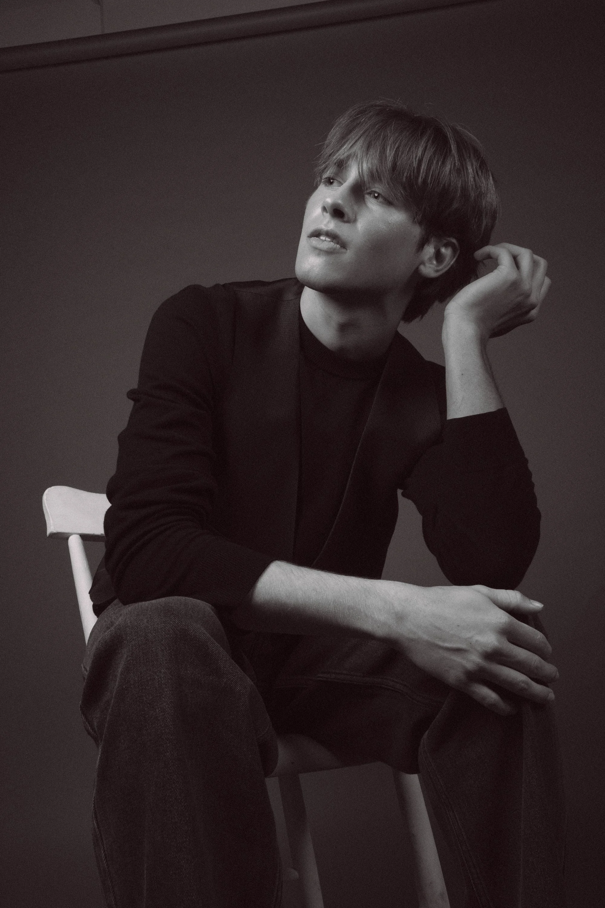 A man in a black long-sleeve shirt sitting on a white chair, leaning on his left arm and gently touching his ear with his right hand, with a thoughtful expression on his face, in a monochrome setting.