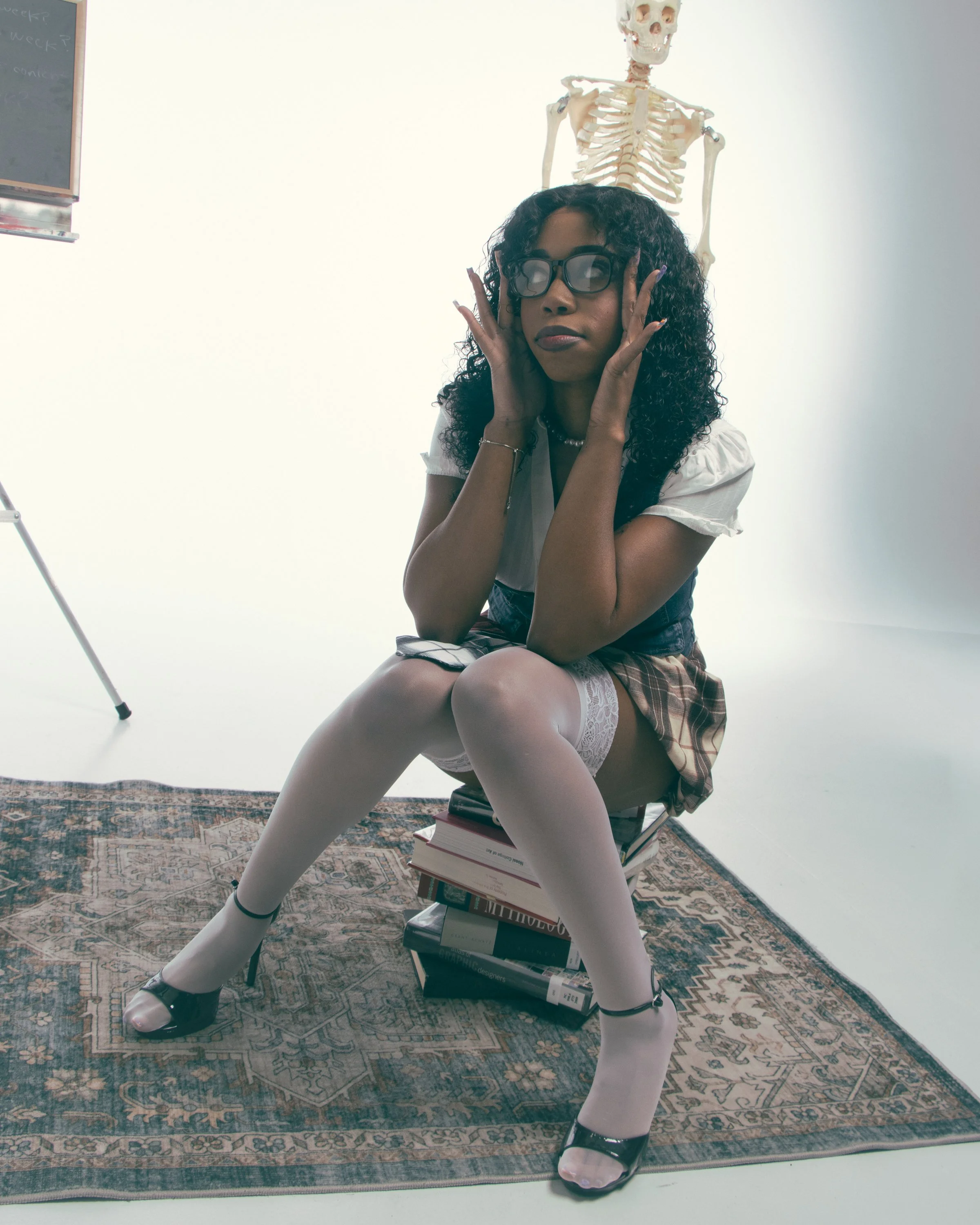 Woman with curly black hair, glasses, white top, plaid skirt, and white thigh-high stockings, sitting on a stack of books, with a skeleton model in the background, on a patterned rug in a studio or classroom setting.