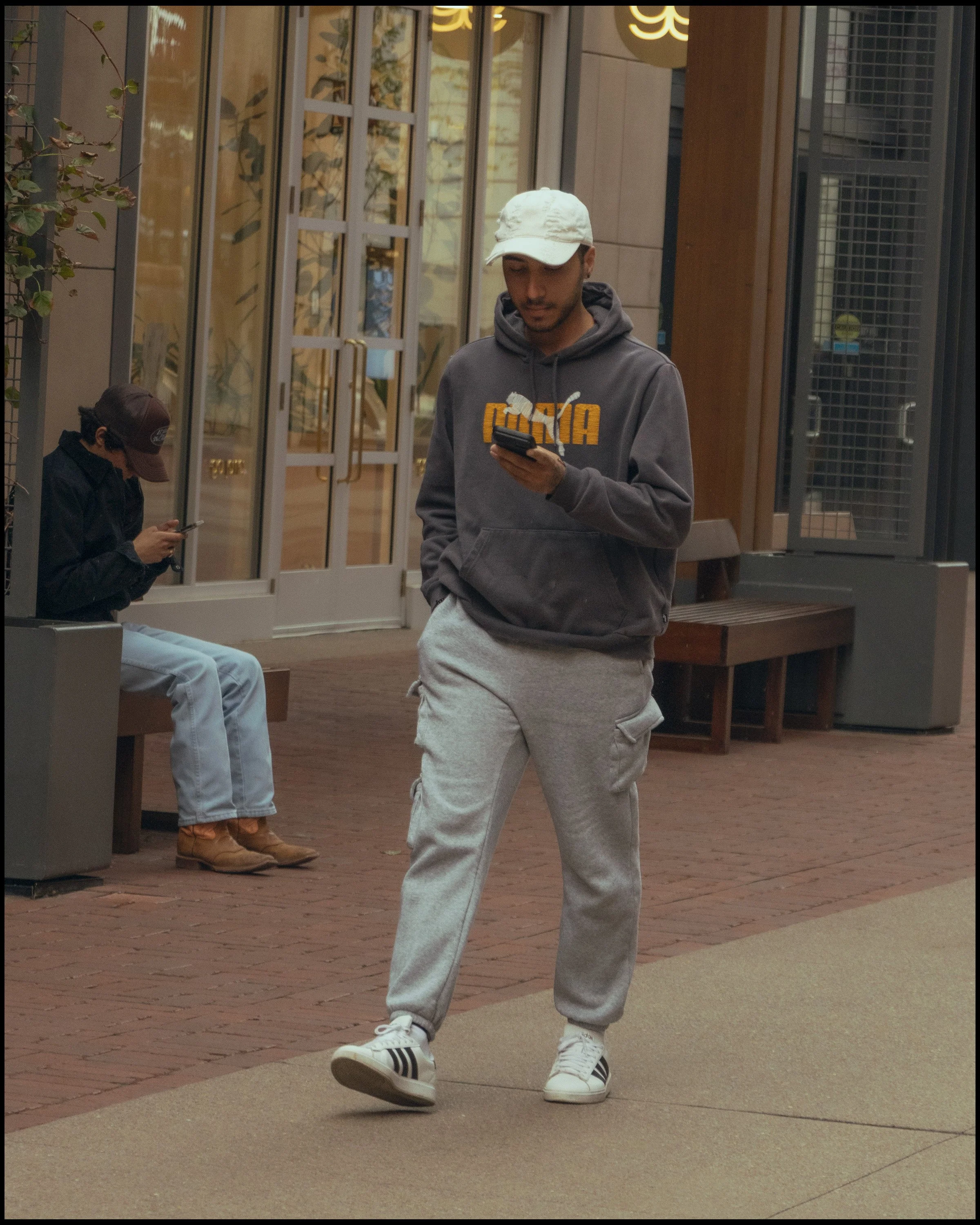 A young man wearing a white cap, gray hoodie with a yellow and white logo, gray sweatpants, and white sneakers with black stripes, walking while looking at his phone in a courtyard area with brick and tile flooring. In the background, another person 